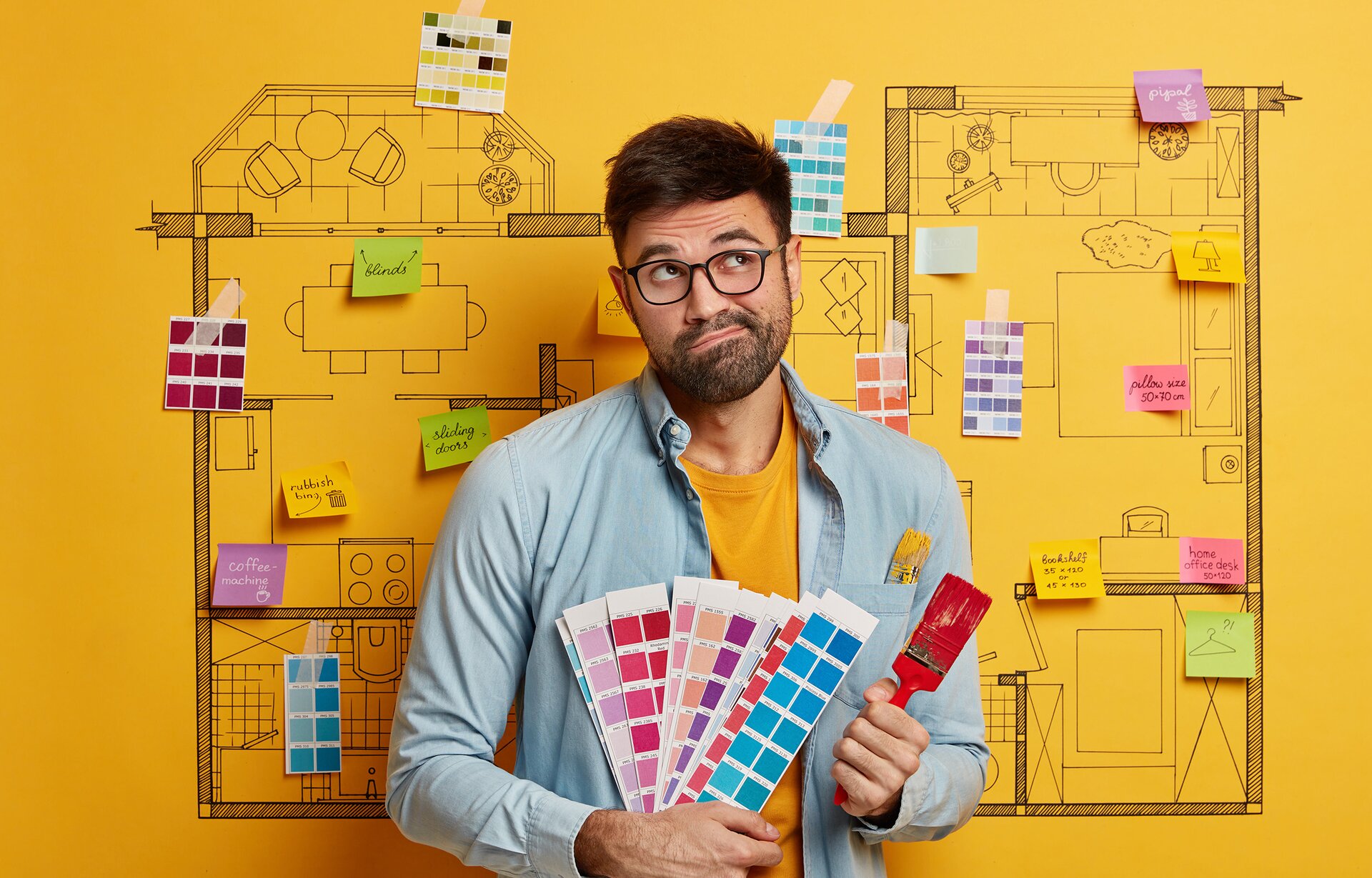 Zdjęcie przedstawia mężczyznę stojącego na tle tablicy zawierającej projekt mieszkania, na której naklejone są różne karteczki. Mężczyzna trzyma w dłoniach znaczniki kolorów oraz pędzel do malowania.
