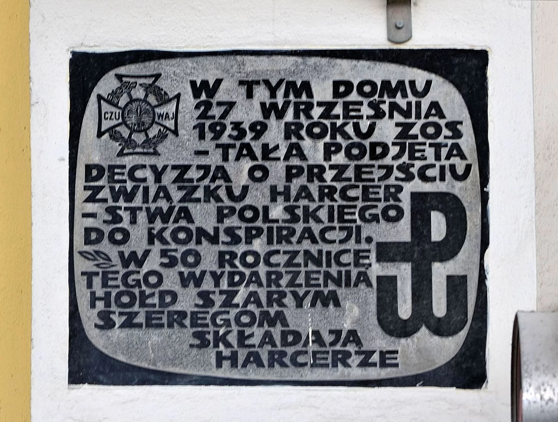 Kliknij, aby powiększyć Tablica. W górnym prawym rogu krzyż opleciony wiankiem z napisem: czuwaj. W dole po prawej stronie widnieje symbol polski walczącej - splecione litery P i W. Na tablicy napis: W tym domu 27 września 1939 roku została podjęta decyzja o przejściu Związku Harcerstwa Polskiego do konspiracji. W 50 rocznicę tego wydarzenia hołd szarym szeregom składają harcerze.