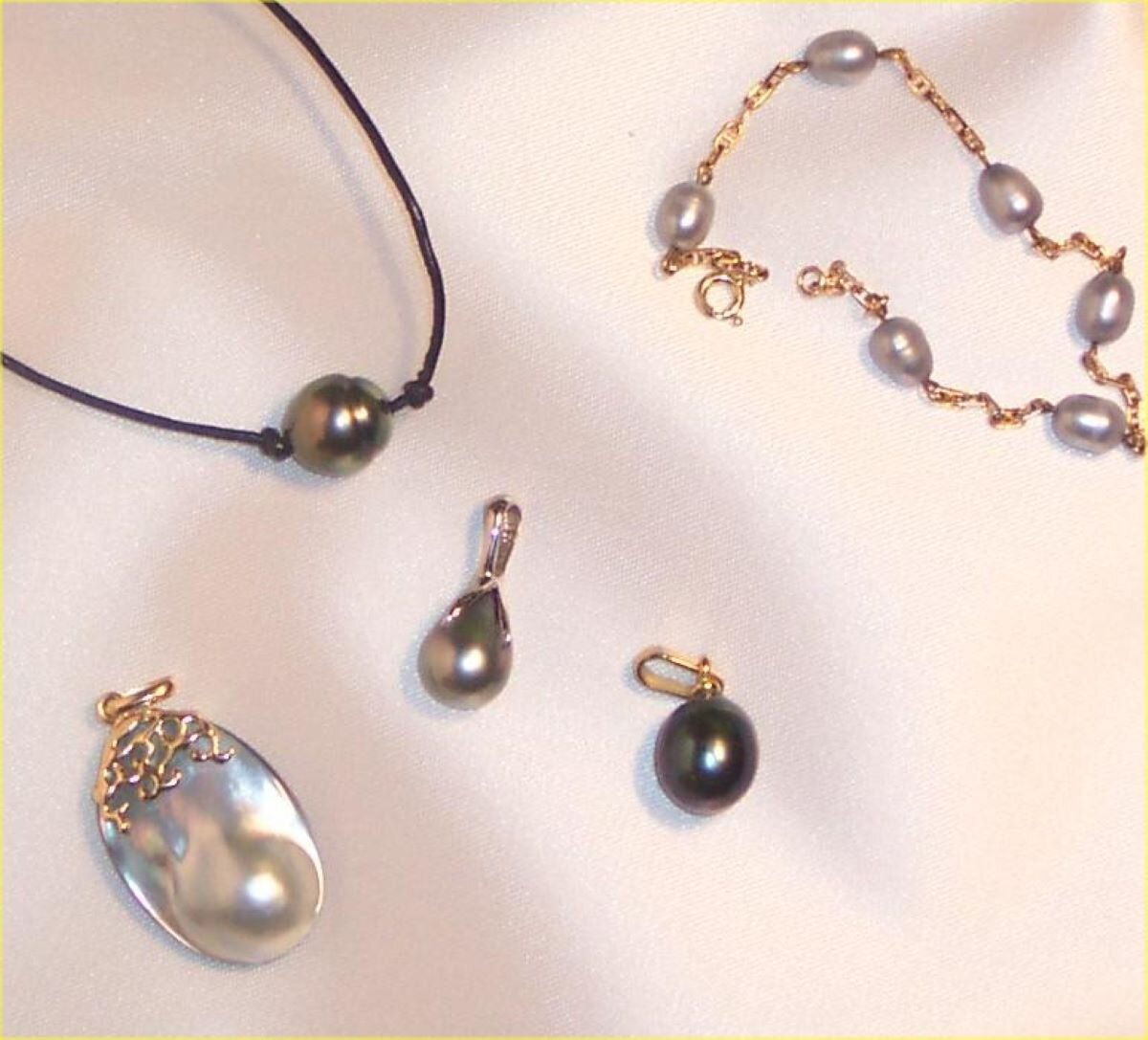 Ilustracja przedstawia zdjęcie wykonane z góry pięciu różnym wytworom biżuteryjnym wykonanym z pereł. Są to: naszyjnik z jedną perłą, złota bransoletka z sześcioma perłami umieszczonymi w jednakowej odległości od siebie, wisior z jasnej perły, kolczyki z ciemnych pereł.