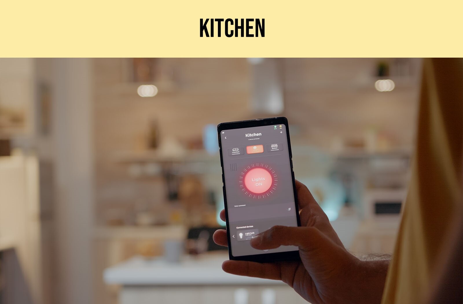 Zdjęcie przedstawia prawą dłoń trzymającą telefon typu smartfon. Na ekranie telefonu widoczne jest czerwone słoneczko z napisem w języku angielskim: Lights ON, powyżej którego widoczne są trzy ikony i napis w języku angielskim: Kitchen. W tle widoczne rozmyte pomieszczenie. U góry ilustracji widoczny jest napis w języku angielskim: KITCHEN.