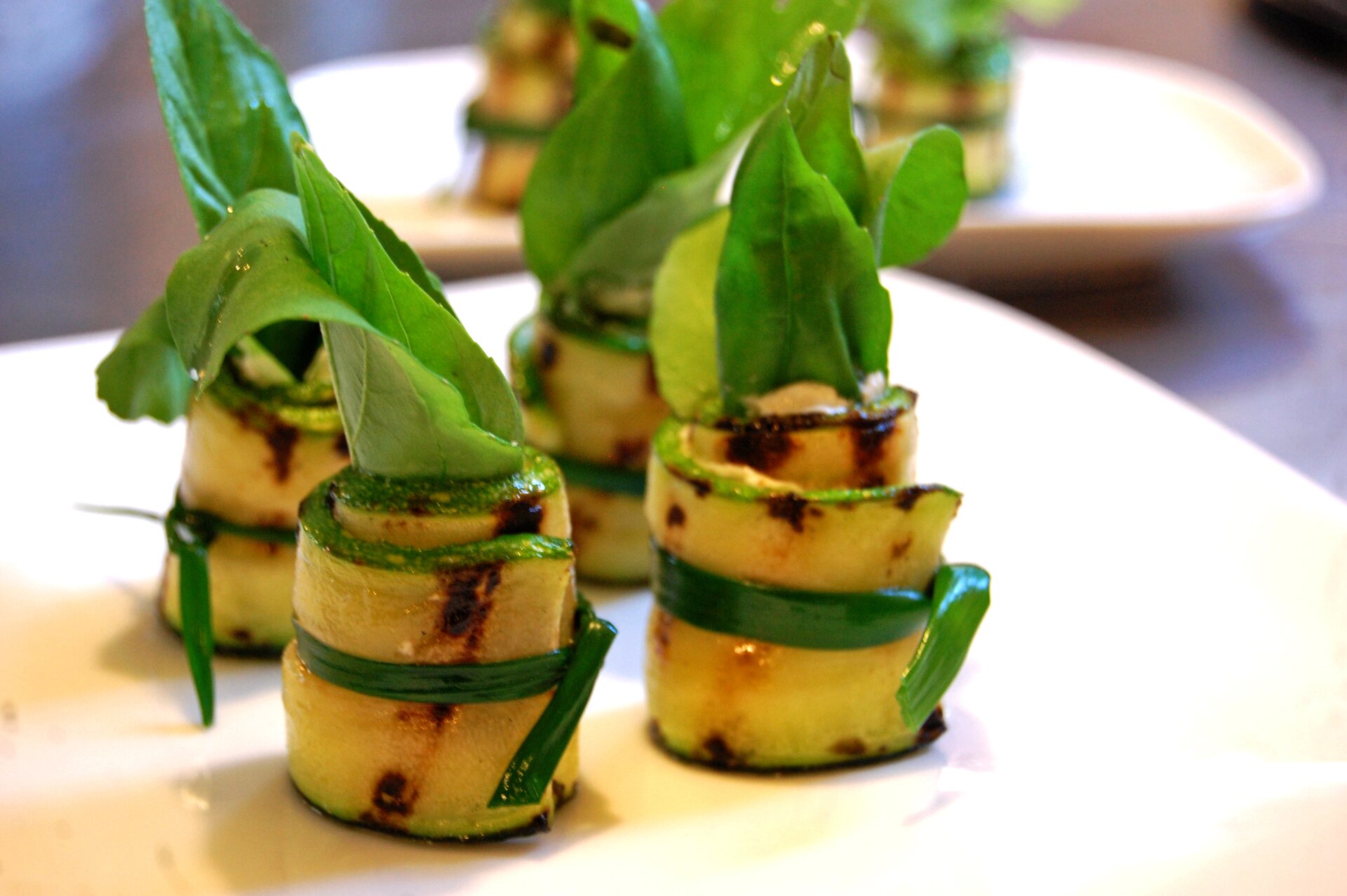 Fotografia przedstawia ustawione na białym talerzu cztery przystawki ze zrolowanych plastrów cukinii, związane szczypiorem lub trawą. Na powierzchni cukinii widać nieregularnie przysmażone pionowe pasy. Od góry z rolek wystają zielone liście sałaty. W tle widoczny jest ciemny blat, a także drugi talerz z podobnym daniem.