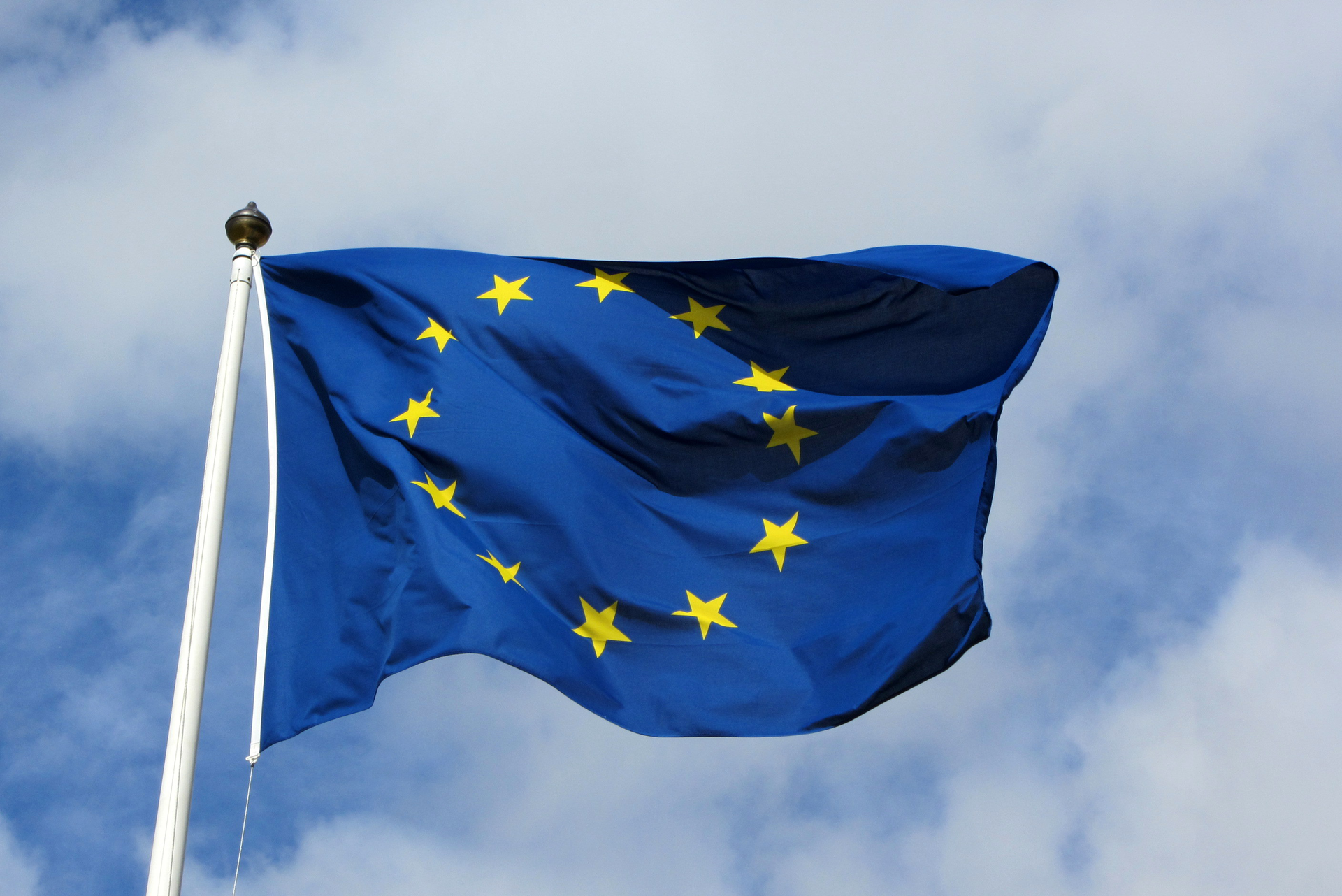 Zdjęcie przedstawia flagę Unii Europejskiej zawieszoną na maszcie. Maszt po lewej stronie zdjęcia. Flaga powiewa skierowana w prawo. Na granatowym tle 12 pięcioramiennych gwiazdek. Gwiazdy ułożone są w okrąg.