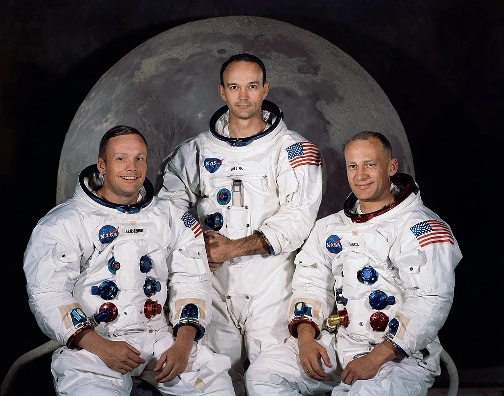 Kliknij, aby powiększyć Na zdjęciu, na tle księżyca pozują trzej astronauci. Mają na sobie białe skafandry, ale ich głowy są odsłonięte. Na skafandrach jest napis NASA, na lewym ramieniu flaga USA oraz na klatce piersiowej sześć otworów do których można coś podłączyć. Astronauci są zadowoleni, uśmiechają się.