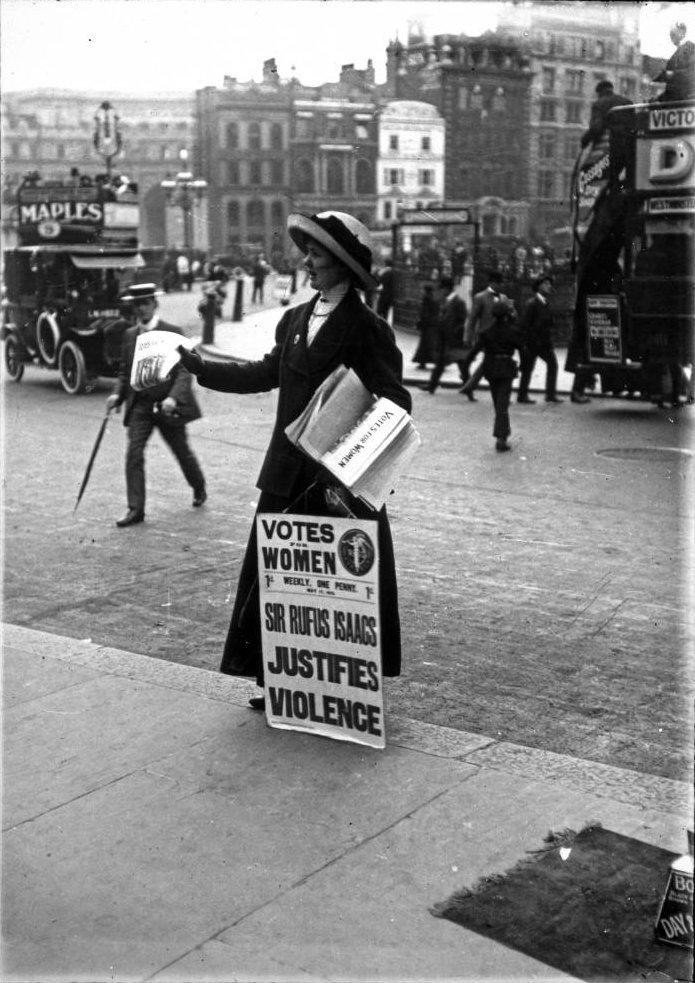 Zdjęcie przedstawia kobietę w długiej sukni i kapeluszu, która stoi na ulicy i rozdaje gazetki. Przed kobietą stoi tabliczka z następującym napisem: Votes for women. Sir Rufus Isaacs justifies violence. W tle znajdują się samochód, przechodnie oraz zabudowania miejskie.