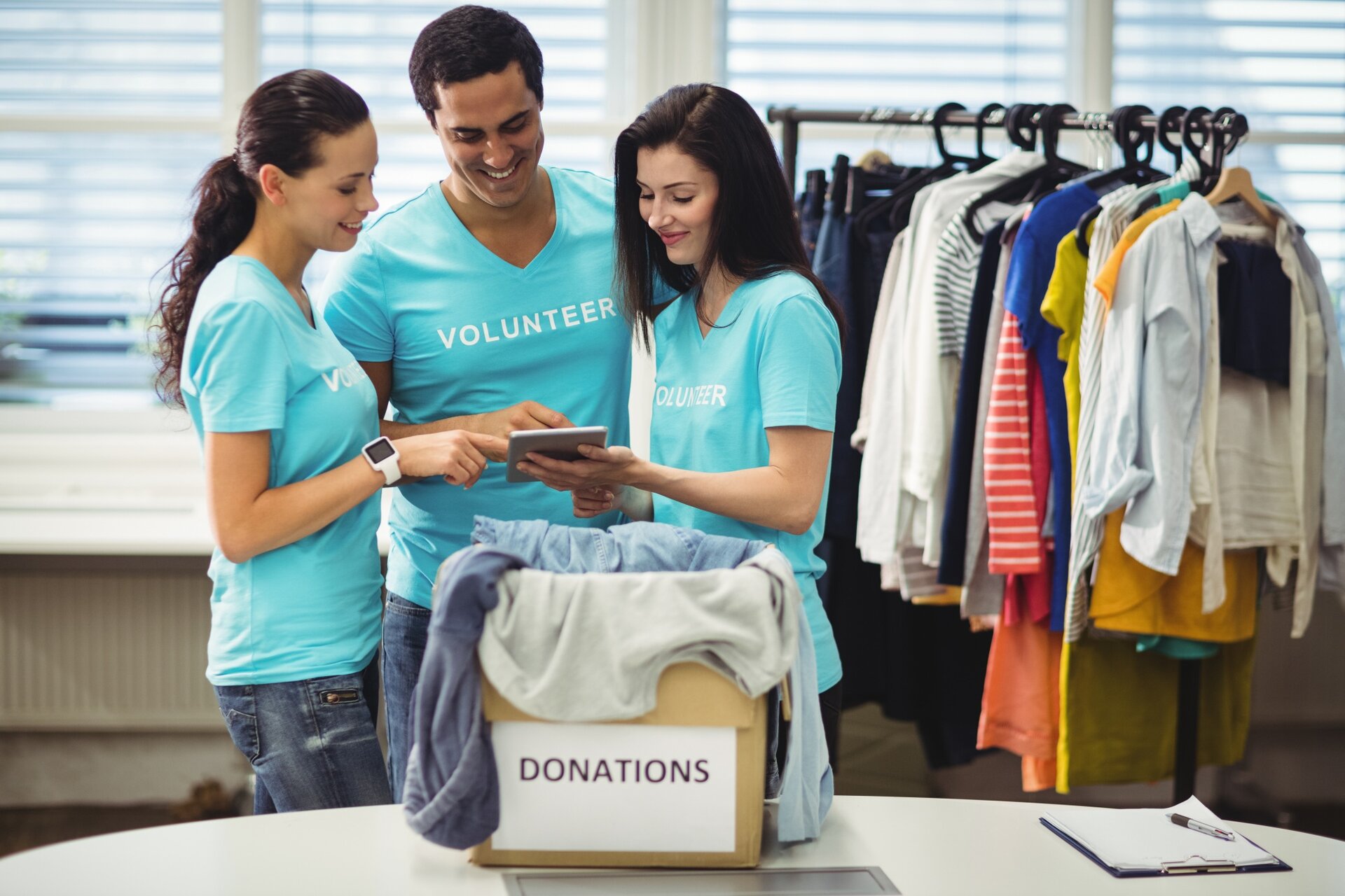 Zdjęcie przedstawia troje młodych ludzi stojących obok siebie. Dwie kobiety, a między nimi mężczyzna. Wszyscy mają na sobie jasnoniebieskie koszulki z napisem "Volunteer". Jedna z kobiet trzyma tablet, w kierunku którego patrzy cała trójka. Na stole, przed nimi stoi pudełko z którego wystają ubrania. Na pudełku jest napis "DONATIONS".