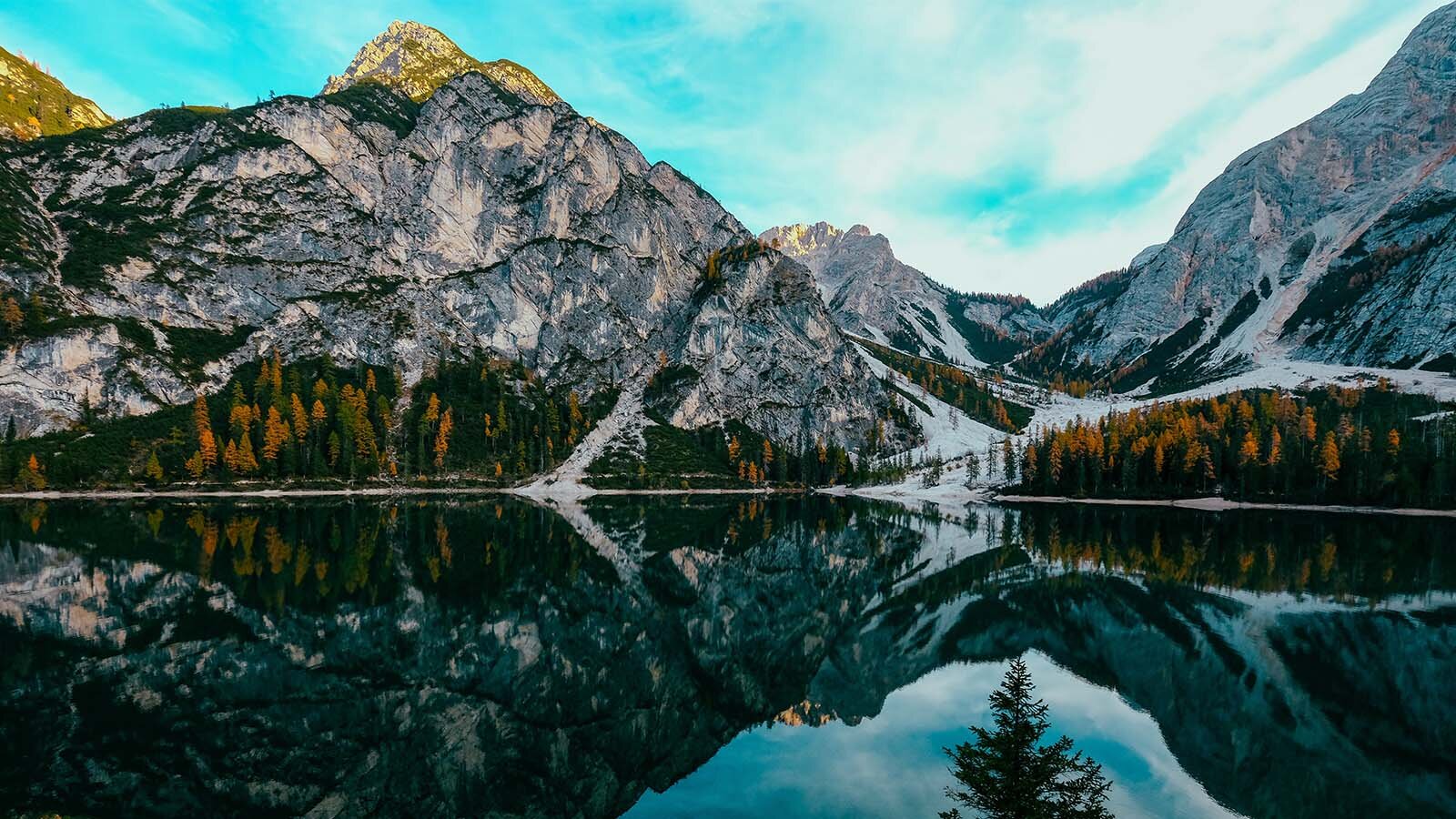 Zdjęcie przedstawia górski zbiornik wodny, którego woda jest intensywnie ciemnoniebieska. W tafli wody odbijają się szczyty skalistych gór w kolorze szarym. W tle widoczne jest skaliste pasmo górskie ze skałami w kolorze biały i szarym, a za nim oświetlony słońcem pojedynczy szczyt. Pośrodku widoczna jest dolina ze schodzącymi po stokach pasmami śniegi. U podnóża gór, nad brzegiem zbiornika, po prawej i lewej stronie, widoczny jest las mieszany, w którym drzewa liściaste mają pożółkłe liście.