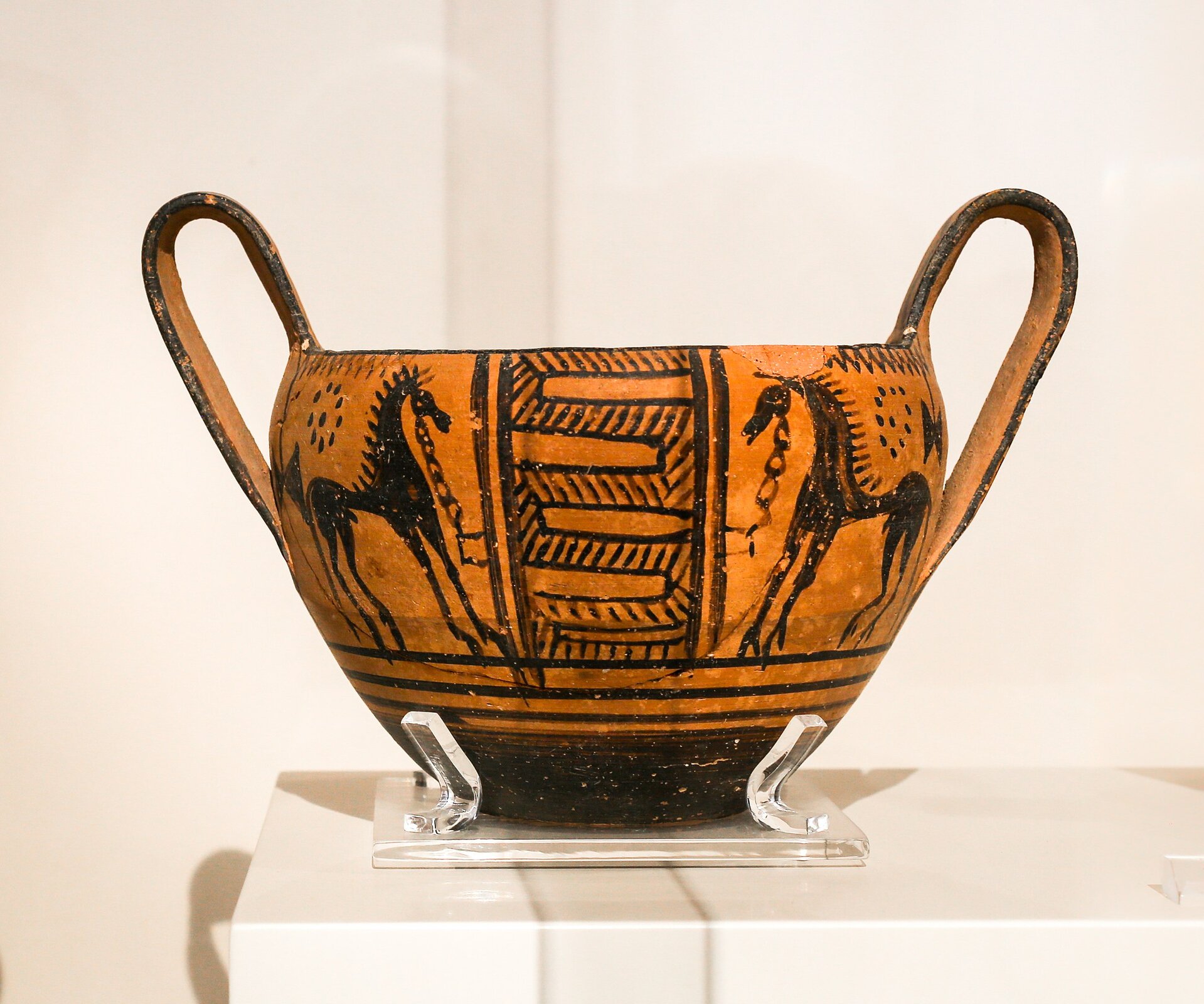 Fotografia autorstwa ArchaiOptix przedstawiający starożytny kantaros, który kształtem przypomina filiżankę lub miskę z dwoma podłużnymi uszami wystającymi wysoko ponad szyjkę naczynia. Naczynie pomalowane zostało dwoma kolorami, czarnym oraz jasno brązowym, na powierzchni umieszczono parę koni stojących naprzeciwko siebie. Pomiędzy końmi stoi fragment płotu. Naczynie zostało namalowane w stylu czarnofigurowym.