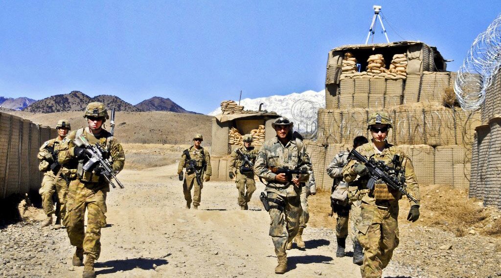 Fotografia przedstawiająca żołnierzy w trakcie misji NATO. Idą ścieżką między zasiekami i prowizorycznym murem. Otacza ich pustynny krajobraz. Mają na sobie mundury polowe w ciepłej, żółtawej tonacji. Na głowach mają hełmy, w rękach niosą broń.