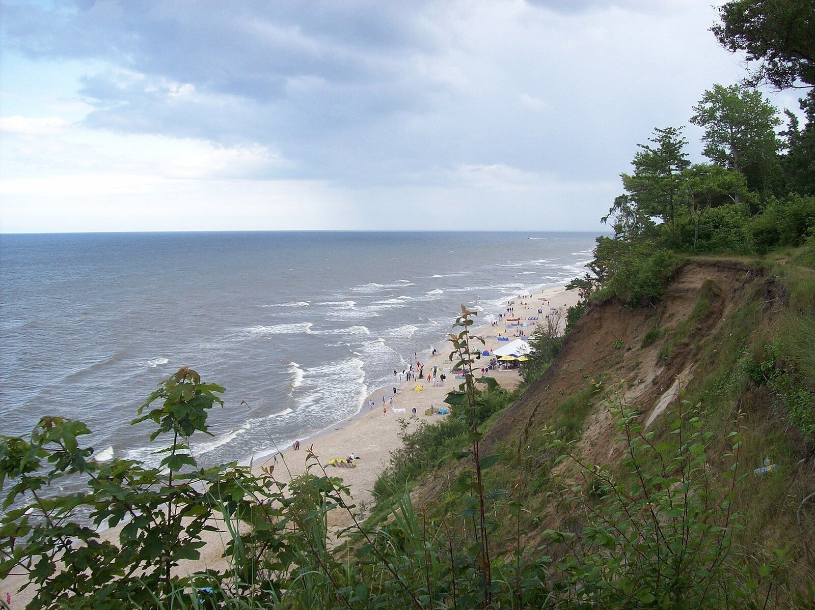 Zdjęcie przedstawia morze z plażą, na której widać ludzi, natomiast po prawej stronie ukazuje się klif porośnięty roślinnością.