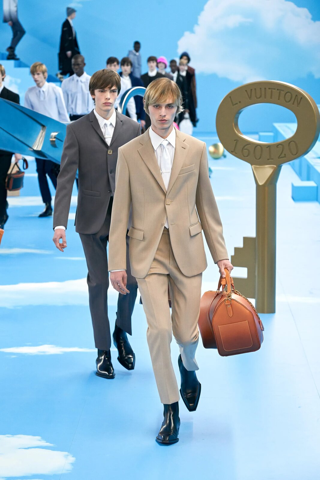 Fotografia przedstawia modeli na pokazie mody. Z przodu widoczni są dwaj młodzi mężczyźni ubrani w beżowy oraz szary garnitur. Pierwszy z nich trzyma w lewej dłoni dużą brązowo‑pomarańczową torbę. Mają na sobie białe koszule oraz krawaty. Z tyłu widoczni są inni modele, również w garniturach. Niektórzy nie mają na sobie marynarek. Po bokach widoczna są elementy dekoracyjne wybiegu, duży klucz ustawiony pionowo, element przypominający duży, drewniany walec, duże drzewo z lewej strony oraz tło i podłoga we wzory przedstawiające chmury.