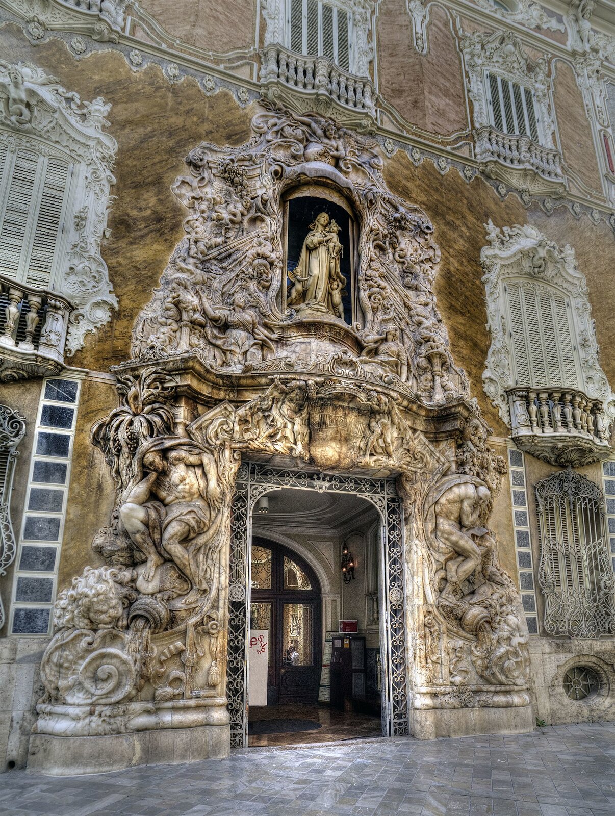 Zdjęcie przedstawia bardzo bogato zdobiony portal. Dookoła prostokątnej wnęki znajduje się mnóstwo rzeźb. Po obu stronach umieszczono postacie. Jedna z nich jest skulona i opiera głowę na dłoni. Druga z nich jest mniejsza i również skulona. Nad wnęką umieszczono wizerunek kobiety w długiej szacie, która trzyma na rękach małe dziecko. Po obu jej stronach stoją małe aniołki. Dookoła tej sceny umieszczono mnóstwo innych rzeźb.