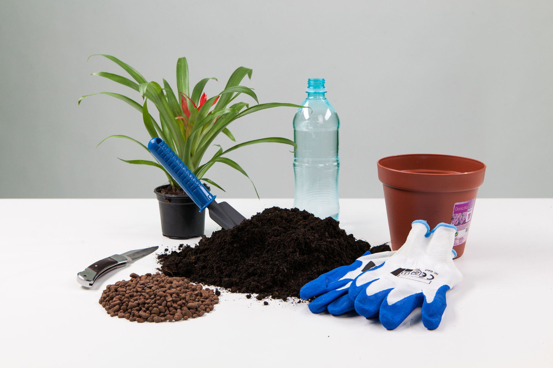 Pokaz slajdów prezentujących przesadzanie rośliny doniczkowej. Slajd 1 – prezentuje przybory potrzebne do przesadzania rośliny: żwir, ziemia, łopatka, nóż, rękawice, nowa doniczka, woda w butelce.