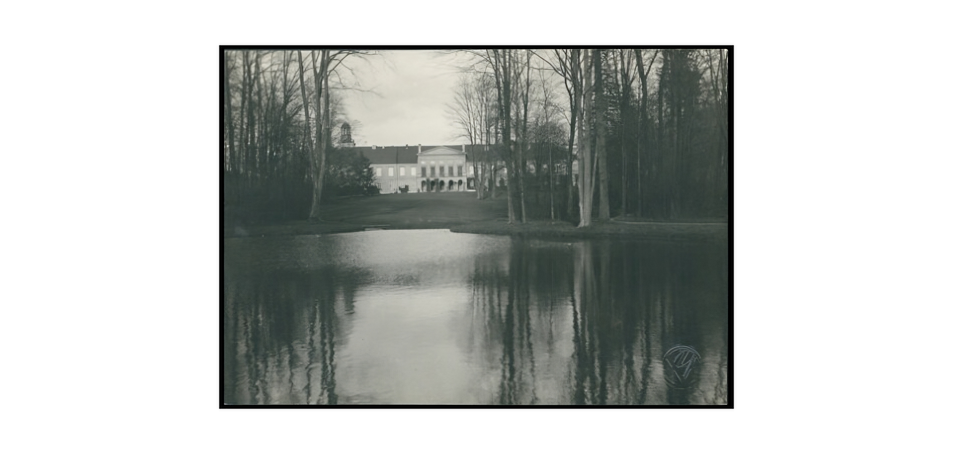 Czarnobiałe zdjęcie przedstawia pałac w Koszęcinie. Na pierwszym planie zbiornik wodny, otoczony bezlistnymi drzewami. Na dalszym planie gmach pałacu. W centrum widoczny szeroki portyk z kolumnadą na parterze, zasłaniający częściowo dwuspadowy dach budynku. Po lewej stronie fotografii widoczny jest fragment wieży zegarowej pałacu. Gmach budynku odbija się częściowo w tafli wody. 