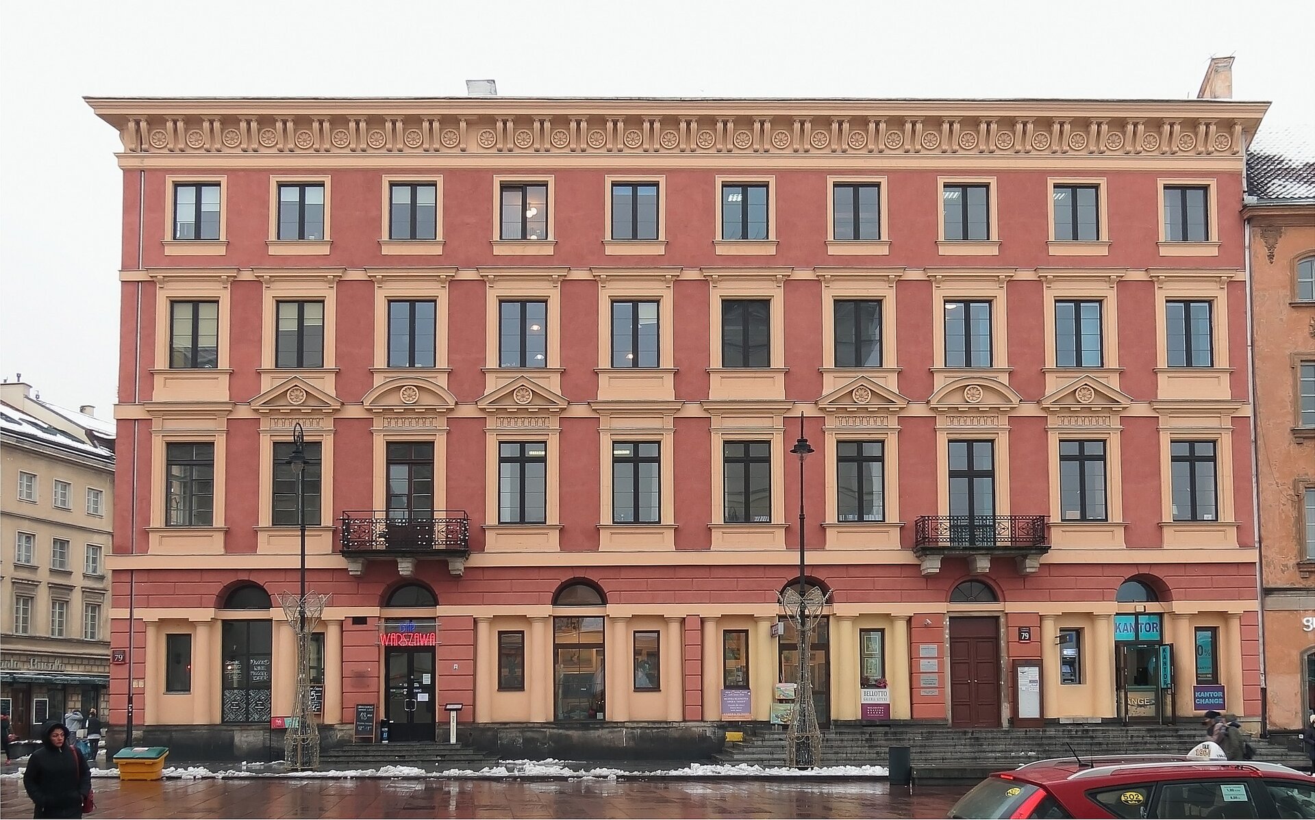 Zdjęcie przedstawia fasadę pierwszego w Polsce nowoczesnego domu handlowego przy Krakowskim Przedmieściu w Warszawie. Pierwotnie budynek był trzykondygnacyjny. Na piętrach znajdowały się mieszkania czynszowe oraz apartamenty czeskich właścicieli kamienicy. Na parterze zaprojektowano specjalne witryny wystawowe. Tworzą je cztery portyki czterokolumnowe nakryte półokrągłym przyczółkiem. Nad nimi znajdują się rzędy obramowanych okien z trójkątnymi i półokrągłymi naczółkami. Pod dachem znajduje się gzyms ozdobiony rzeźbiarską dekoracją.