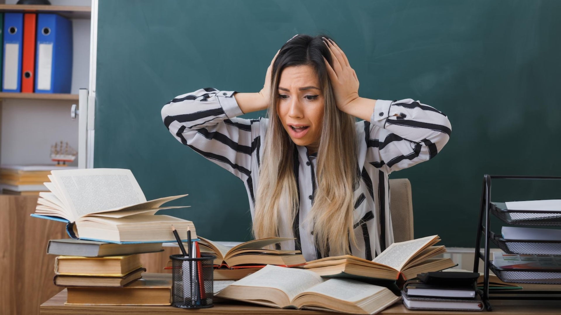Zdjęcie przedstawia kobietę z długimi włosami siedzącą przy biurku wśród dużej ilości książek. Kobieta obiema rękami trzyma się za głowę. Za kobietą znajduje się tablica szkolna.