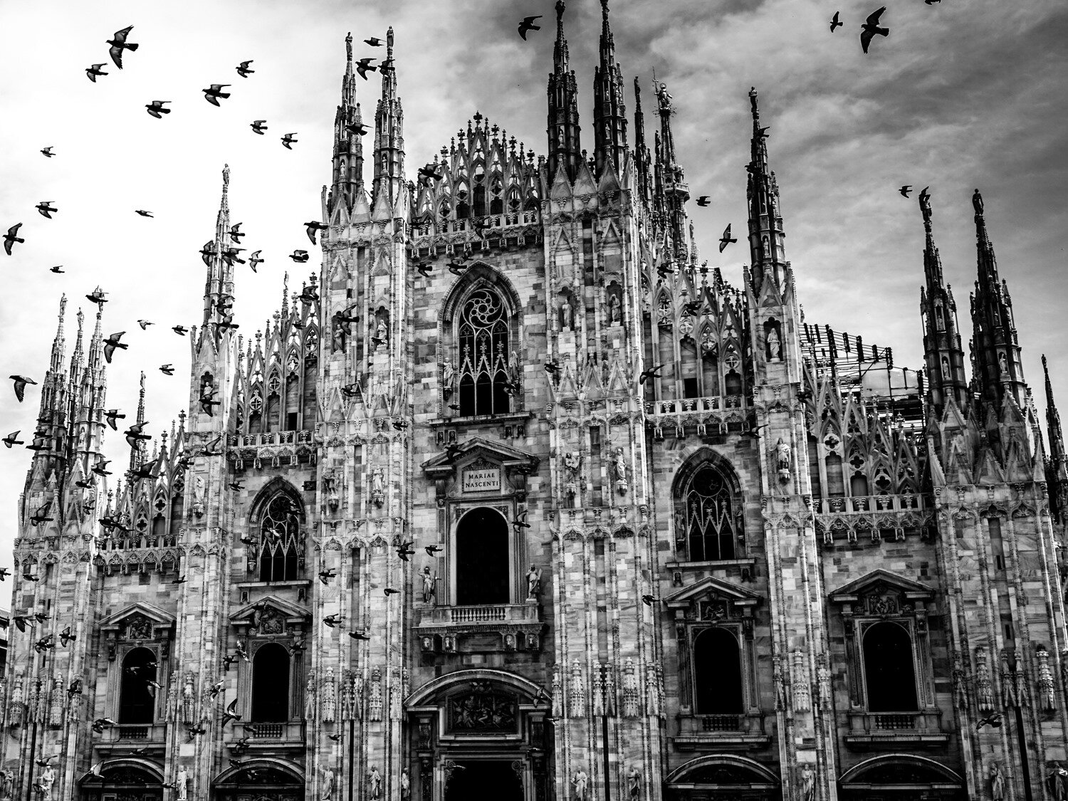 Czarno‑białe zdjęcie przedstawia trójkątną, strzelistą fasadę ogromnej kamiennej katedry. Widoczne są strzeliste przypory, od najniższych po zewnętrznych stronach fasady do coraz wyższych pośrodku. Pomiędzy nimi widoczne są wejścia i wysokie okna wypełnione witrażami  i zwieńczone w górnej części plecionką i ostrym łukiem. Wzdłuż brzegów fasady widoczne są ażurowe detale architektoniczne.