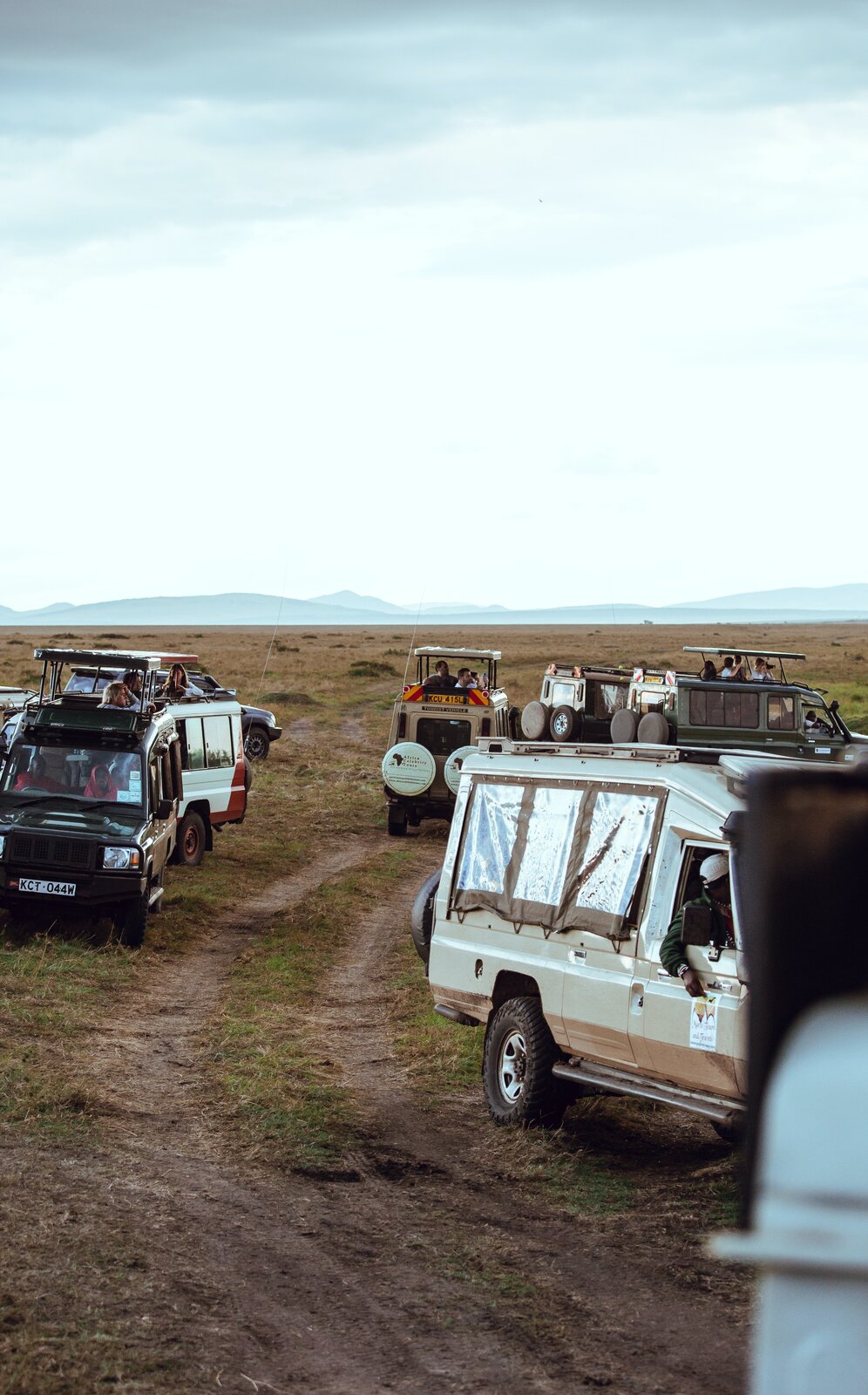 Pośrodku fotografii droga i samochody terenowe. Rozległy, płaski teren porośnięty trawą. W oddali rozciągające się wzniesienia. 