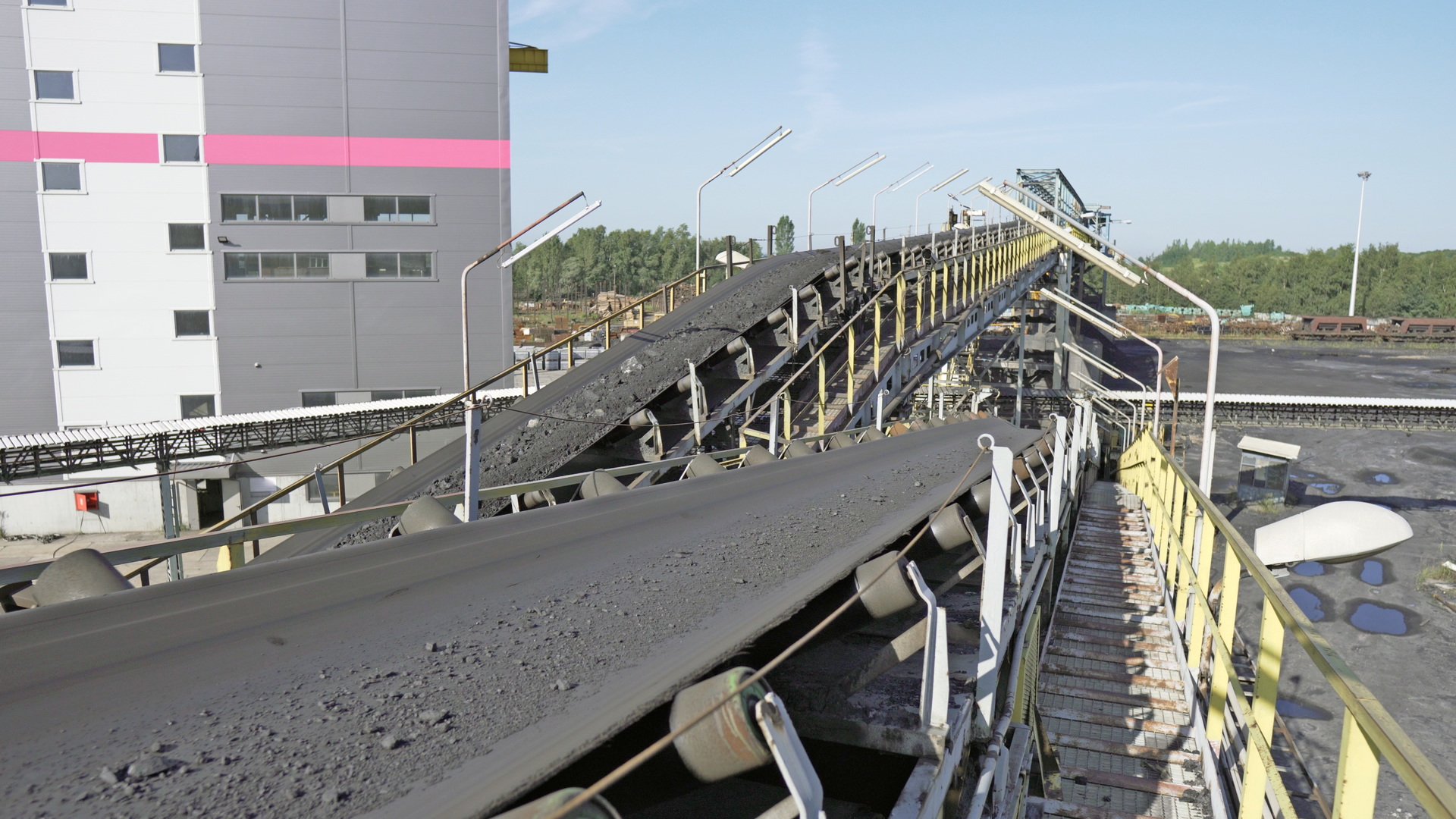 Na zdjęciu widoczne są dwa przenośniki taśmowe. Na taśmie przenośników transportowana jest niewielka ilość węgla. Taśmy osadzone są na walcowatych rolkach. W tle widać budynek przemysłowe