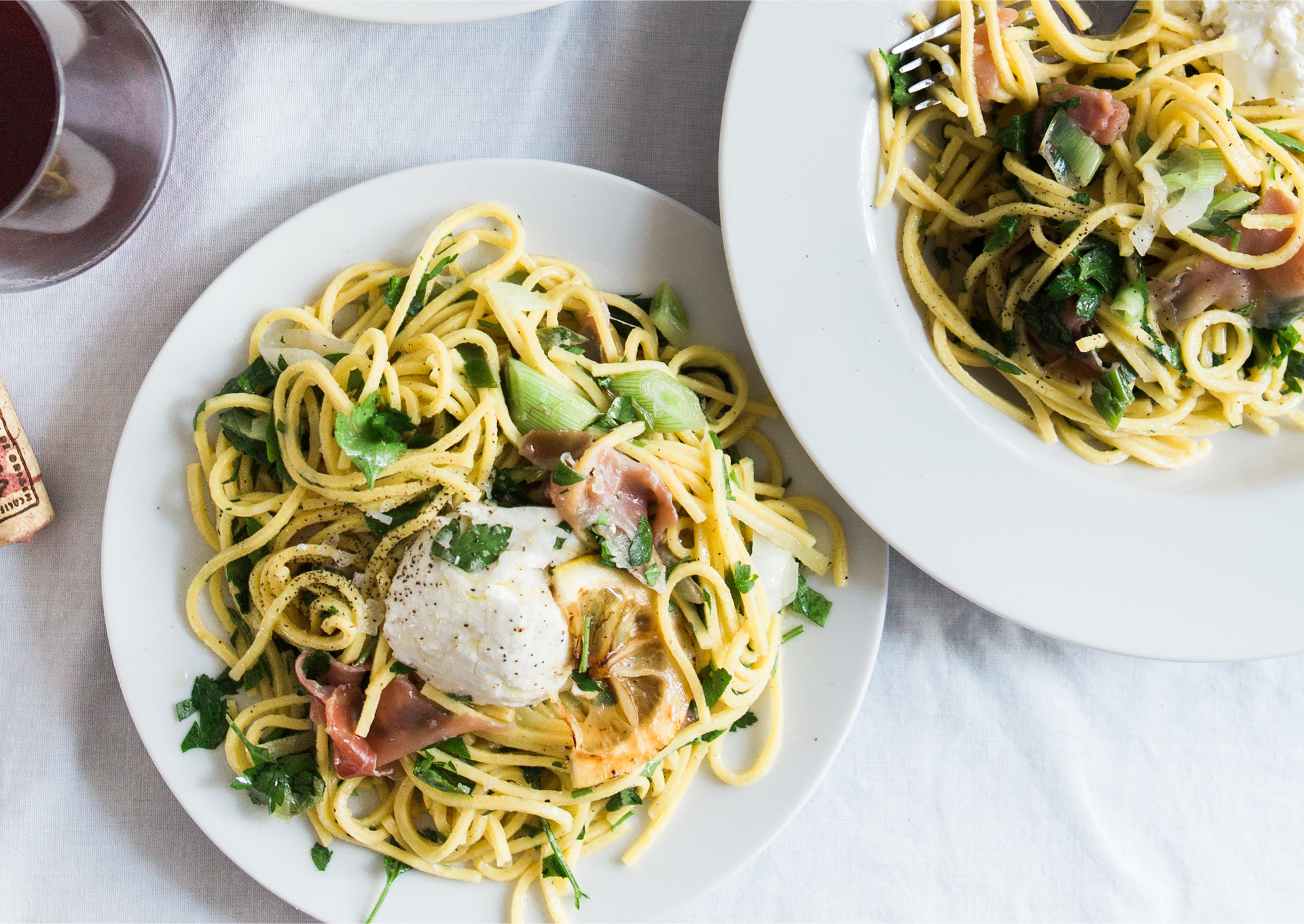Zdjęcie, zrobione z góry, przedstawia stół przykryty białym obrusem, na nim dwa talerze ze spaghetti. Wśród makaronu widać liście różnych ziół, warzyw i długo dojrzewającą włoską szynkę. Na wierzchu jest kleks śmietany.