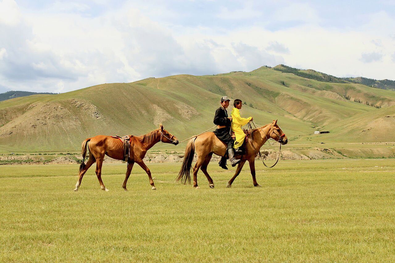 Fotografia barwna przedstawiająca step. Na fotografii płaski pokryty trawą teren. Dwie osoby jadą razem na jednym koniu prowadząc za sobą drugiego konia. W tle porośnięte niską roślinnością wzgórza.