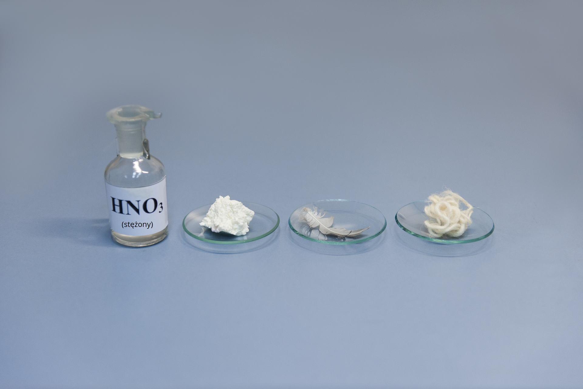Pierwsze zdjęcie w galerii. Na ilustracji prezentowana jest szklana butelka z etykietką i napisem H N O indeks dolny, trzy, koniec indeksu dolnego stojąca na niebieskim podłożu. Po jej prawej stronie, na blacie laboratoryjnym, stoją w szeregu trzy szkiełka zegarkowe, na których położono kolejno: kawałek białego sera, ptasie pióro i zwitek owczej wełny.