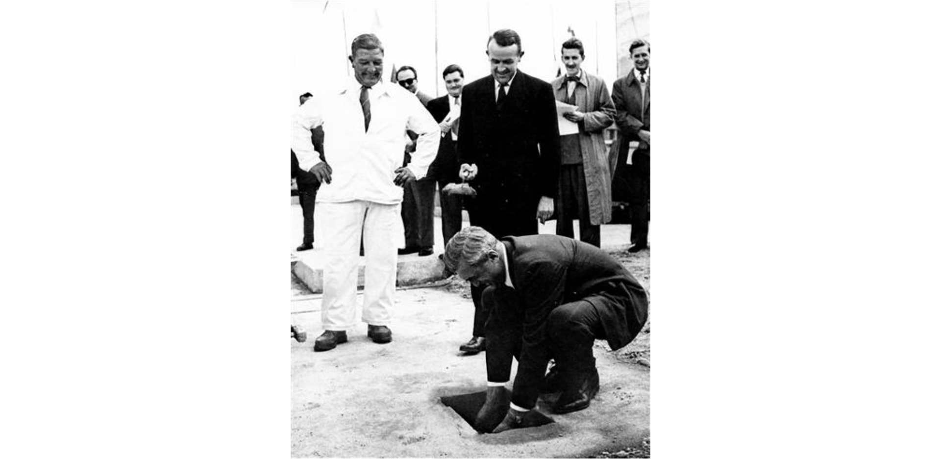 Czarnobiałe zdjęcie. Widoczna jest na nim grupa ludzi. Jeden z nich kuca i ma dłonie włożone w wykopaną w ziemi dziurę.