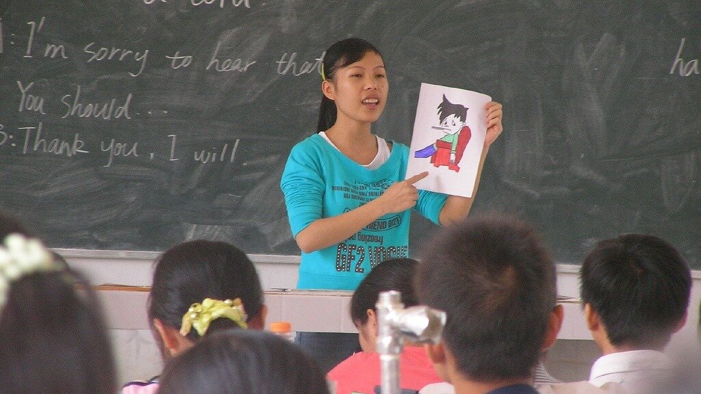 Zdjęcie barwne przedstawiające sytuację w klasie szkolnej, prawdopodobnie w którymś z krajów azjatyckich. Widać tyłem obrócone głowy uczniów, chłopców i dziewcząt, oraz postać nauczycielki, która wskazuje palcem trzymaną w drugiej ręce ilustrację. W tle znajduje się duża szkolna tablica kredowa i kilka zdań: I'm sorry to hear that... You should... Thank you, I will.