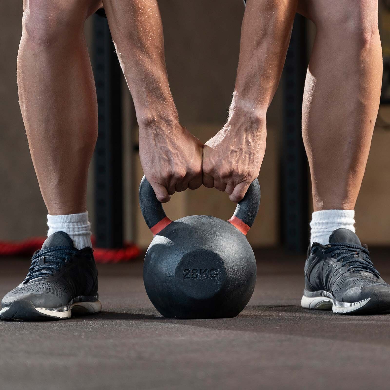 Zdjęcie ukazuje szeroko rozstawione nogi mężczyzny w obuwiu sportowym. Między nogami jest ustawiony ciężar w postaci kuli z górnym uchwytem, o wadze 28 kg. Na uchwycie są zaciśnięte dwie dłonie. Transkrypcja znajduje się poniżej