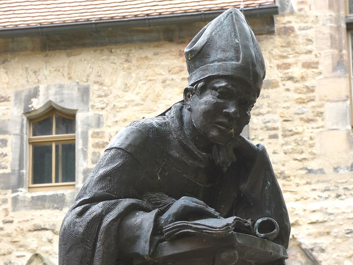 Ilustracja przedstawia współczesną rzeźbę przedstawiającą Thietmara. Na głowie ma czapkę biskupią, w rękach trzyma księgę. Za nim widoczne są mury zamku.