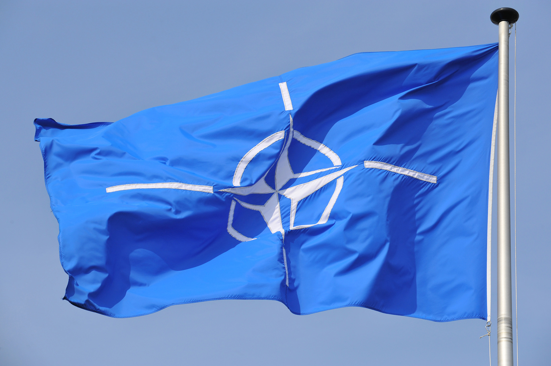 Zdjęcie przedstawia flagę NATO zawieszoną na maszcie. Maszt po prawej stronie. Flaga unosi się i jest skierowana w lewo. Na błękitnym tle znajduje się biały pierścień. W środku pierścienia czteroramienna gwiazda. To tak zwana róża kompasowa. Ramiona gwiazdy są zakończone białym, wąskim paskiem.