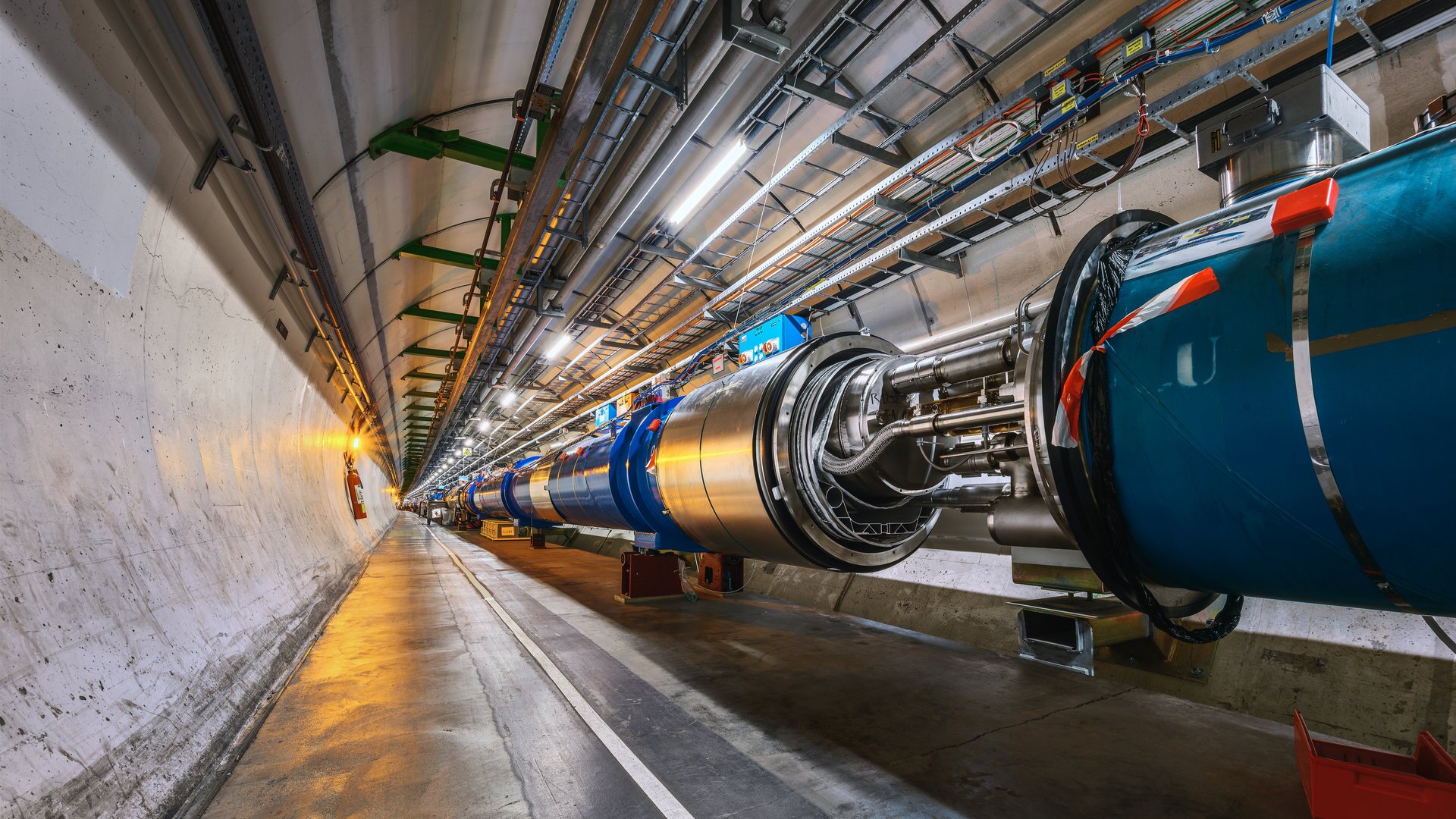 Rys. 1. Zdjęcie poglądowe przedstawia fragment wielkiego zderzacza hadronów (LHC). Wygląda to jak betonowy korytarz z wielką metalową kolorową rurą z podłączeniami.