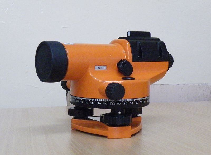 Na zdjęciu znajduje się niwelator automatyczny. Ustawiony jest na trójbocznej podstawce. W dolnej części umieszczona jest miarka. Urządzenie wyposażone jest w instrument optyczny, które przybiera kształt walca i stanowi główną część niwelatora.