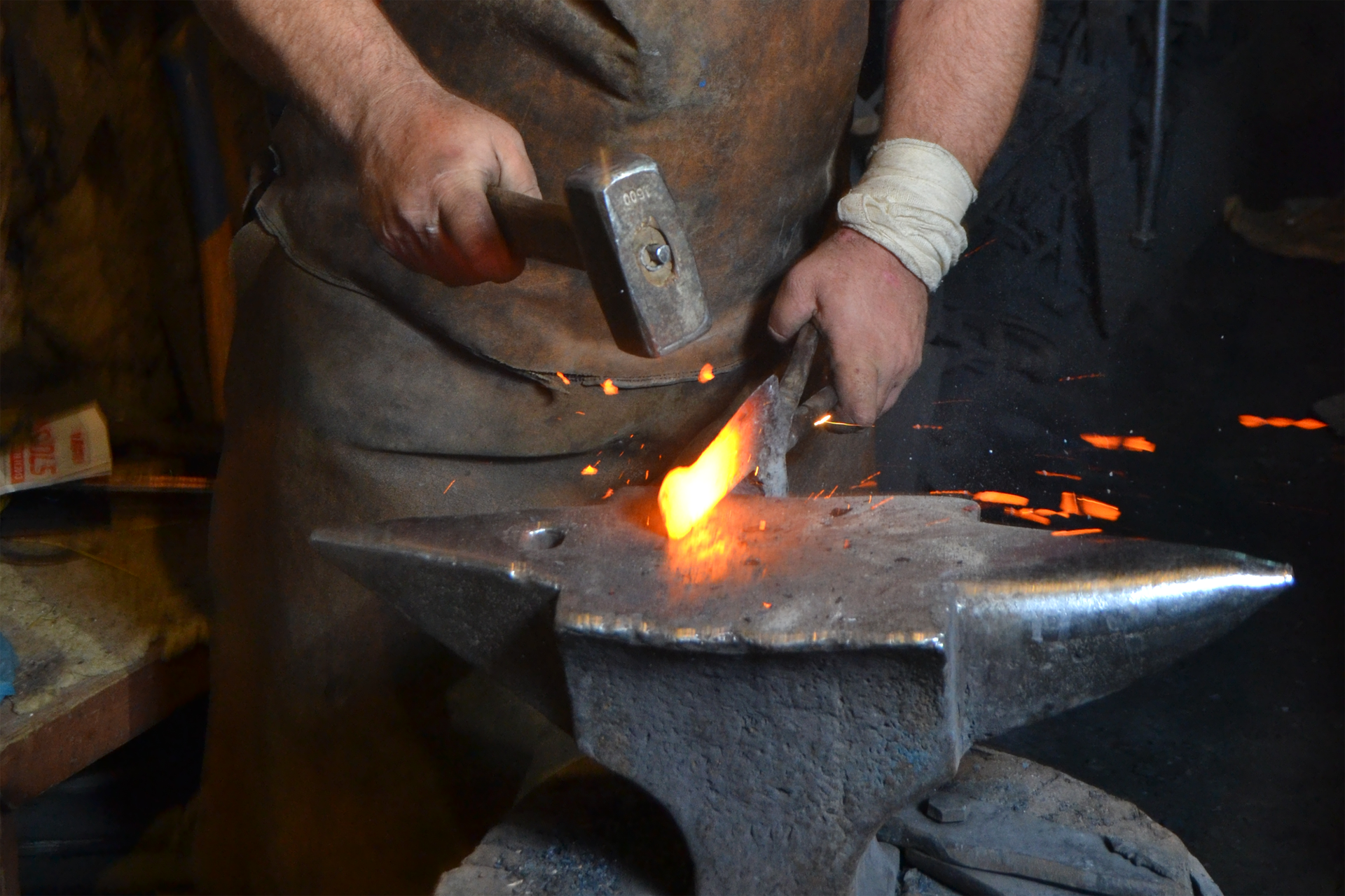 Fotografia prezentuje scenę w kuźni. Kowal kuje rozgrzany do czerwoności metal. W jednej ręce trzyma szczypce, którymi stabilizuje metal, w drugiej ręce trzyma młotek, którym uderza w metal. Metal opiera o kowadło.