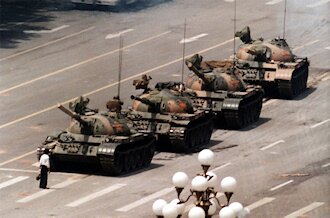Zdjęcie przedstawia plac, po którym jadzie kolumna czterech czołgów. Przed pierwszym z nich stoi człowiek.