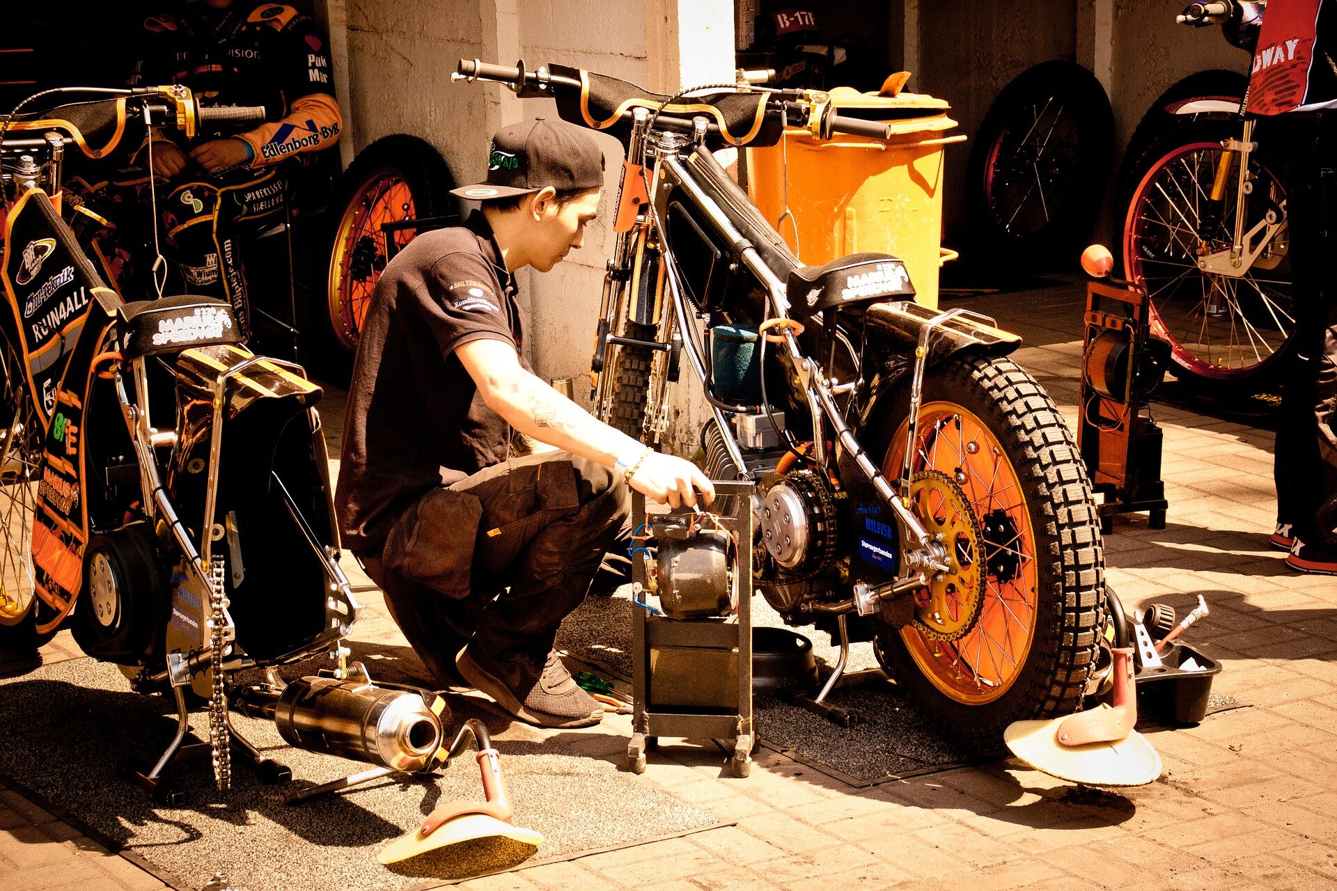 Zdjęcie ukazuje młodego mężczyznę naprawiającego motocykl. Pojazd ma opony z wyraźnym bieżnikiem. 