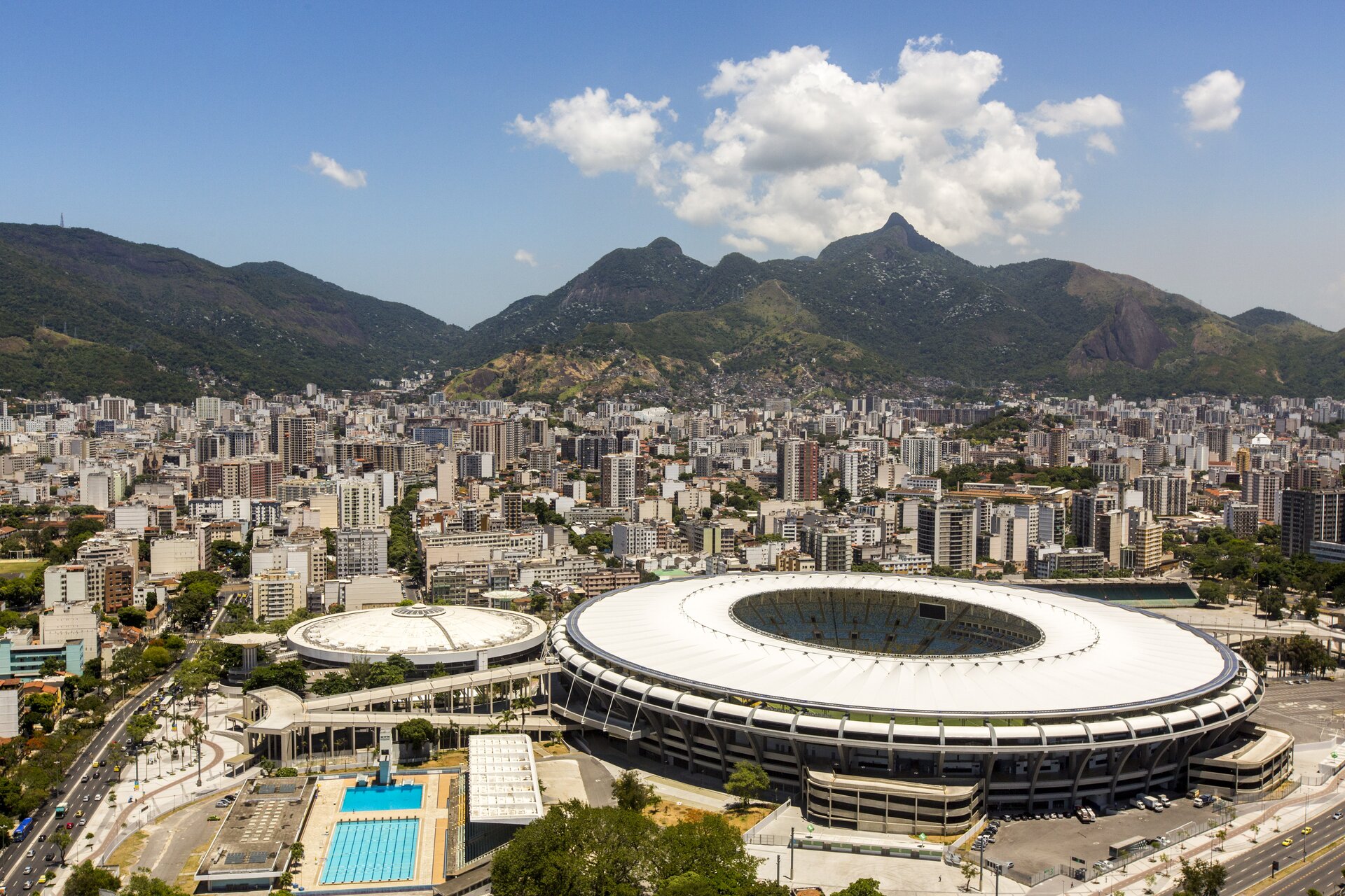 Na zdjęciu miasto u stóp gór, nowoczesne wysokie budynki, na pierwszym planie kompleks zabudowań sportowych z okrągłym stadionem i basenami.