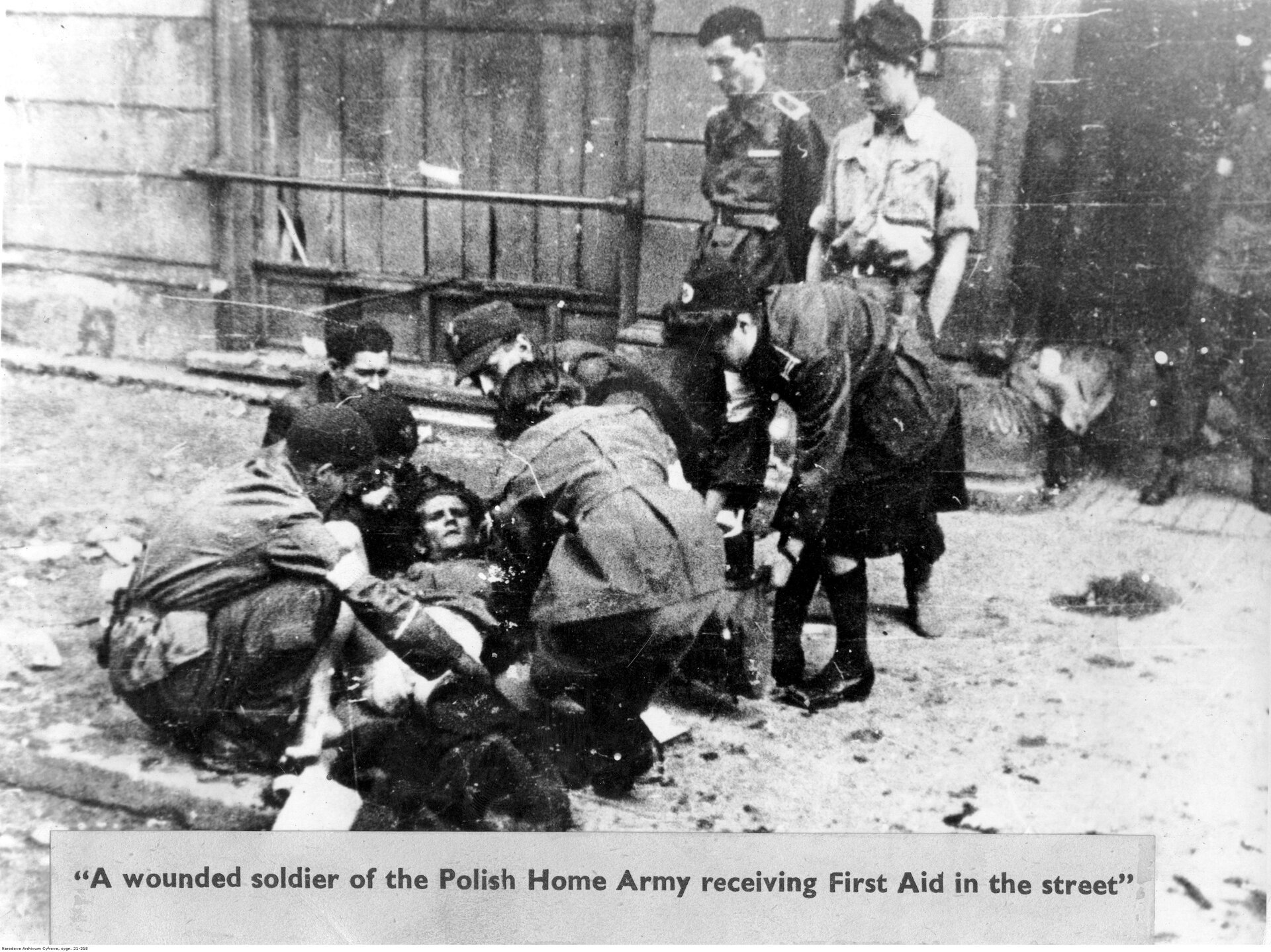 Zdjęcie przedstawia grupę osób pochylających się nad rannym żołnierzem leżącym na chodniku. Usiłują mu pomóc. Jest to młody mężczyzna z włosami zaczesanymi do tyłu. Ma wysokie czoło i zmarszczone brwi. Obok stoją trzy inne osoby. Patrzą na rannego. W głębi, po prawej stronie ukazana jest brama kamienicy. Stoi tu umundurowany mężczyzna. W głębi, pośrodku ukazana jest witryna sklepowa zasłonięta deskami. Pod spodem znajduje się podpis:  A wounded soldier of the Polish Home Army receiving First Aid in the street.