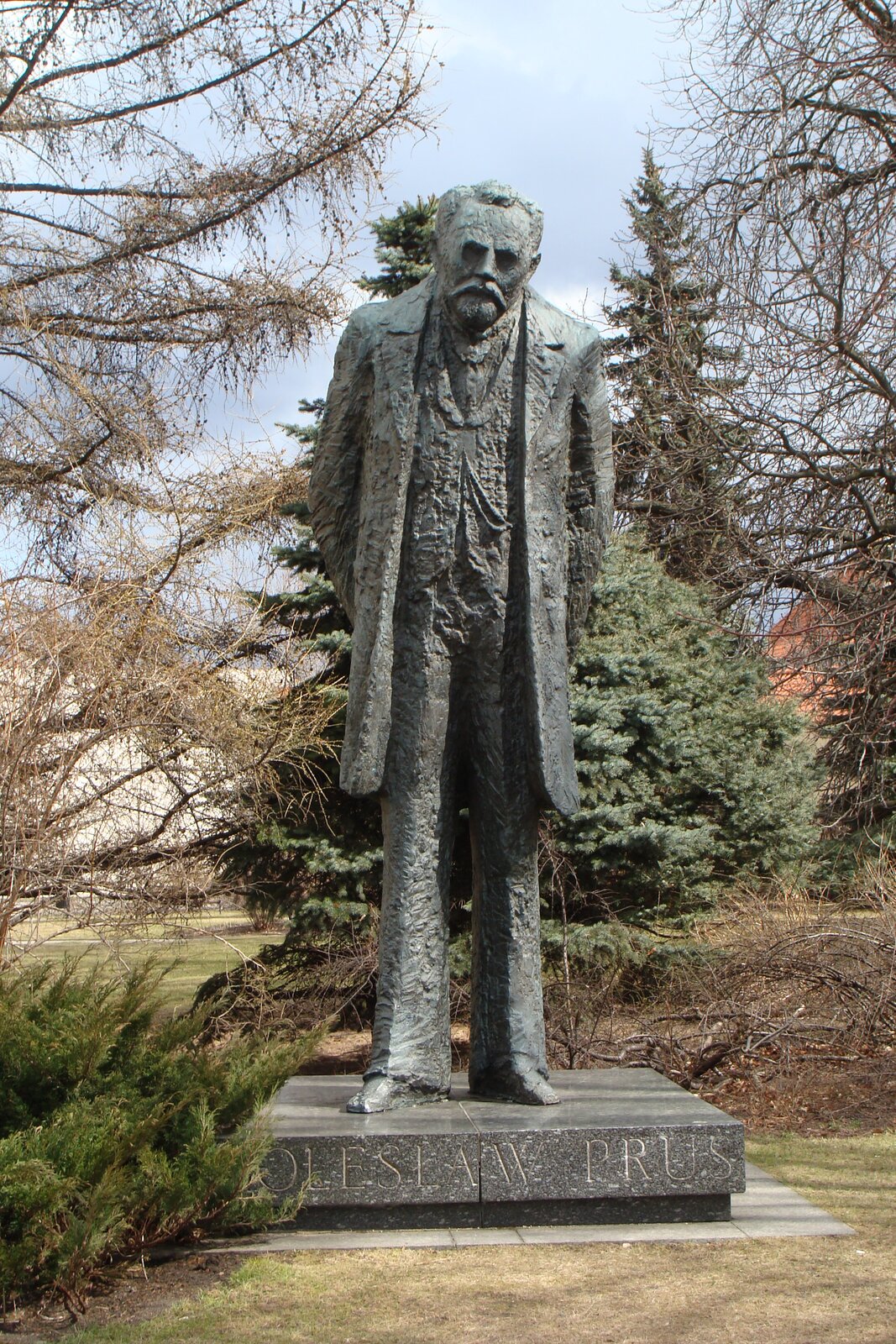 Zdjęcie przedstawia pomnik starszego mężczyzny. Jest lekko pochylony, ma krótkie włosy, krótką bródkę i spiczaste wąsy. Ubrany jest w surdut. Pomnik stoi na podeście z napisem Bolesław Prus. Wokół rosną drzewa.