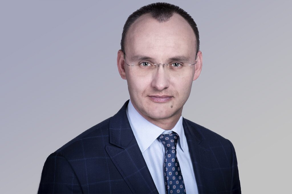 Na zdjęciu mężczyzna w wieku średnim. Z bardzo krótką fryzurą. Mocno widoczne zakola, przedzielone fragmentem włosów. Lekko uśmiechnięta twarz. Nosi okulary. Ubrany w garnitur. Pod garniturem widoczna koszula z krawatem.