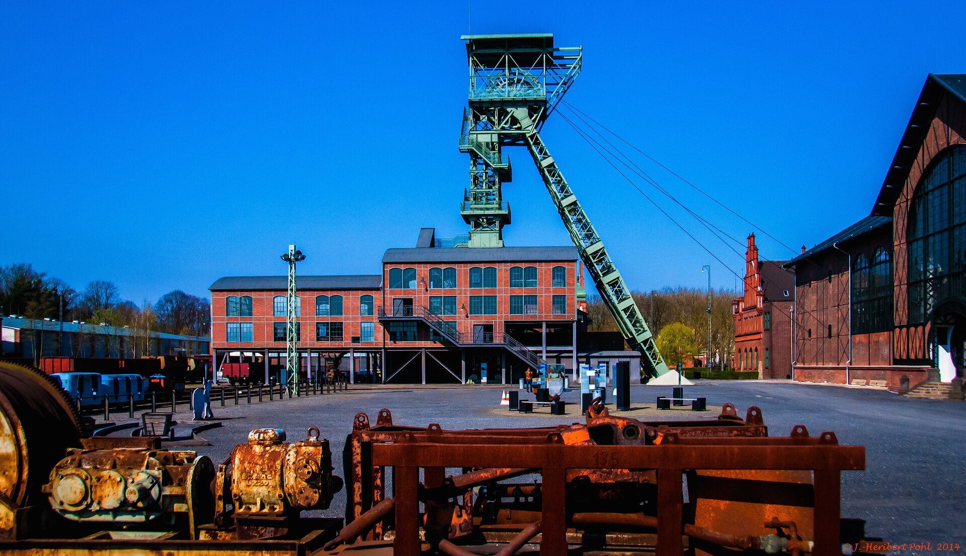 Na zdjęciu Muzeum Zeche Zollern w Dortmundzie, Na pierwszym planie od dołu zdjęcia wystaje część zardzewiałej maszyny, w tle budynek z czerwonej cegły a za nim szyb kopalniany w zielonym kolorze.