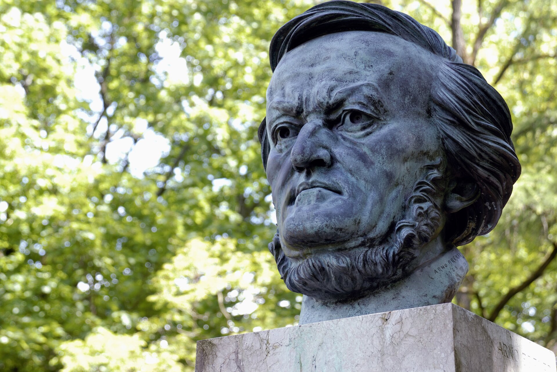 Ilustracja przedstawia pomnik Ryszarda Wagnera. Na zdjęciu widoczna jest odlana z brązu głowa kompozytora, umieszczona na cokole. Rzeźba przedstawia mężczyznę w średnim wieku z lekko dłuższymi włosami i brodą.