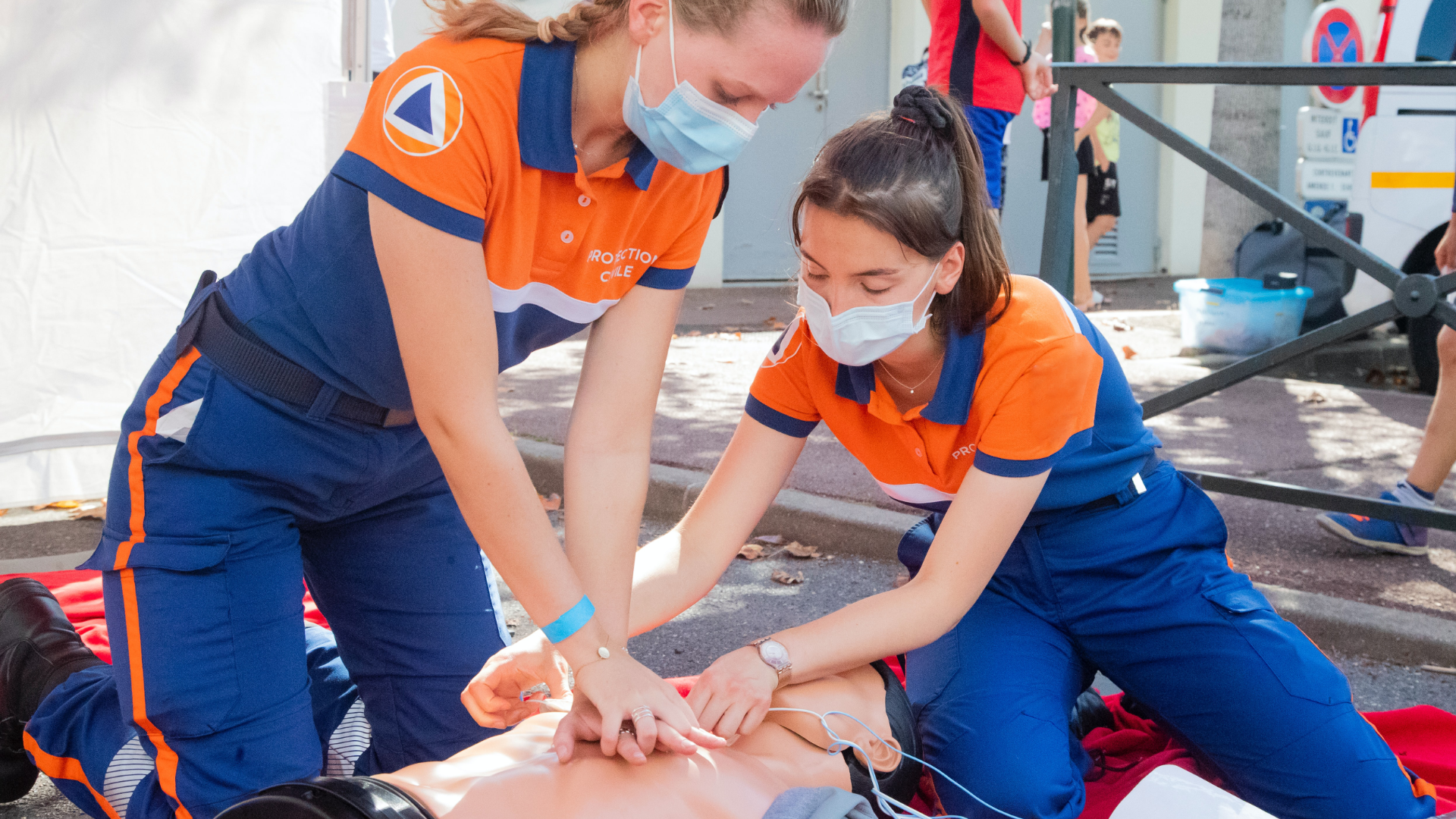 Zdjęcie przedstawia dwie kobiety, które są ubrane w stroje ratowników medycznych. Na twarzach mają maseczki ochronne. Kobiety prezentują na fantomie jak udzielić pierwszej pomocy. Jedna z kobiet wykonuje masaż serca na manekinie.