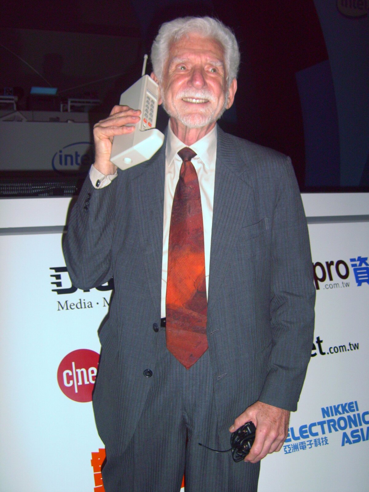 Ilustracja przedstawiająca Martina Coopera z prototypem telefonu komórkowego. Trzyma on w ręku przy uchu duży bezprzewodowy telefon z antenką. W drugiej ręce ma ładowarkę do tego telefonu. W tle znajduje się ścianka z logami różnych firm komputerowych.