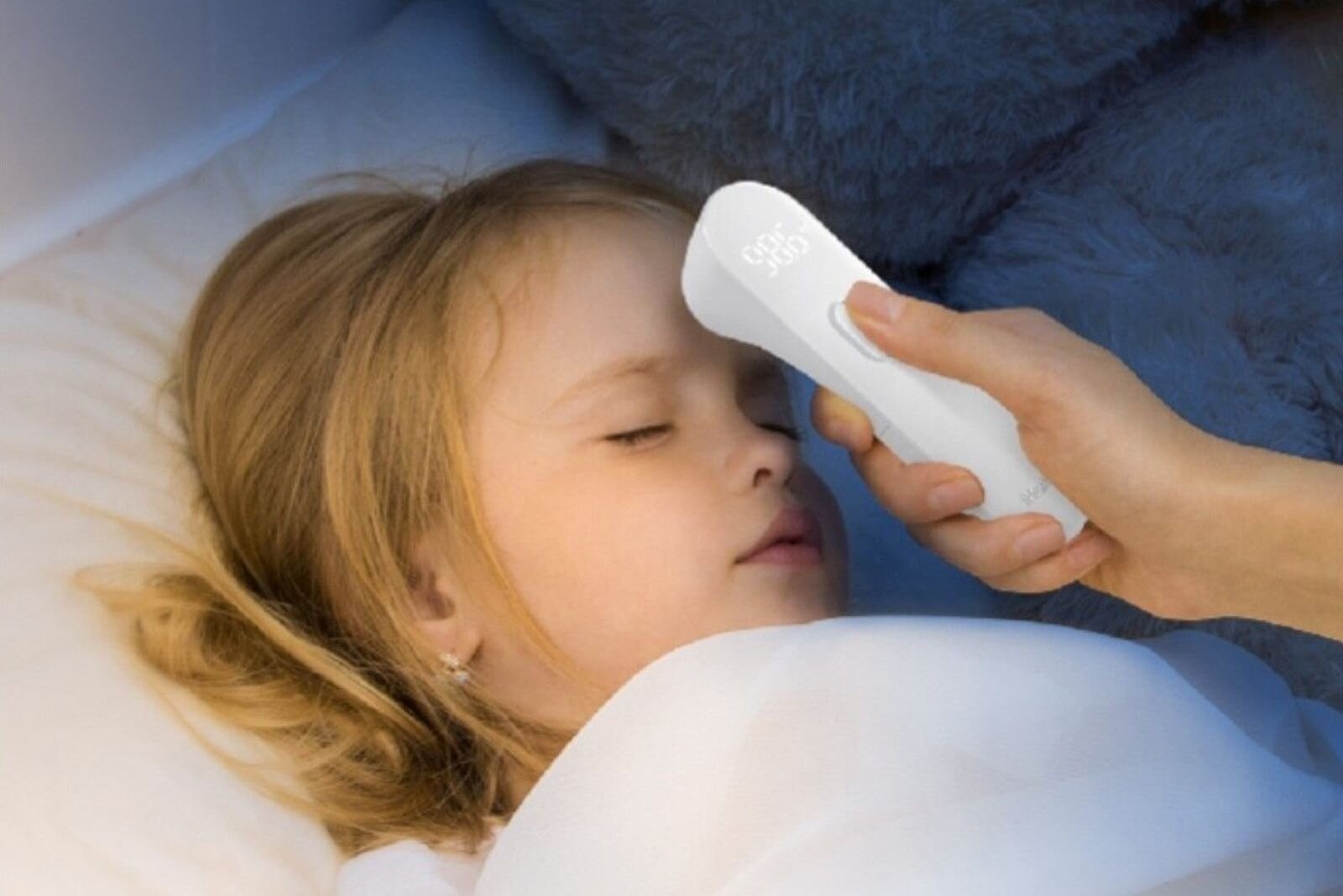 Zdjęcie przedstawia lekarski termometr bezdotykowy w użyciu. Widać dłoń trzymającą taki termometr. Ma on podłużny kształt z szerszą końcówką. Ta końcówka zbliżana jest do czoła leżącej w łóżku dziewczynki.