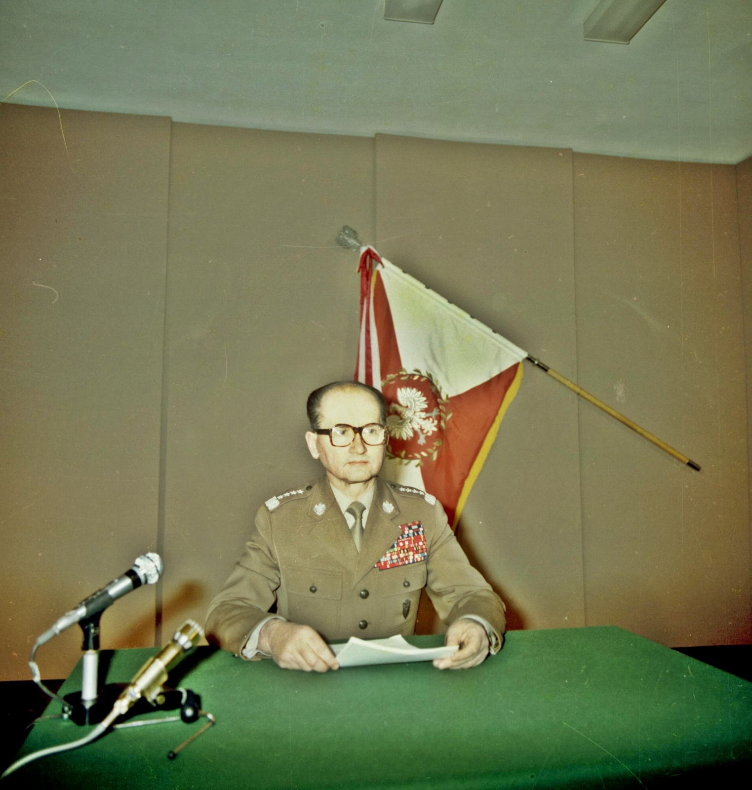 Zdjęcie przedstawia dojrzałego mężczyznę w mundurze generalskim. Mężczyzna siedzi, trzyma kartkę, ma założone okulary, jest łysiejący. Na stole z zielonym obrusem stoją mikrofony. Za mężczyzną wisi biało-czerwony sztandar z białym orłem.