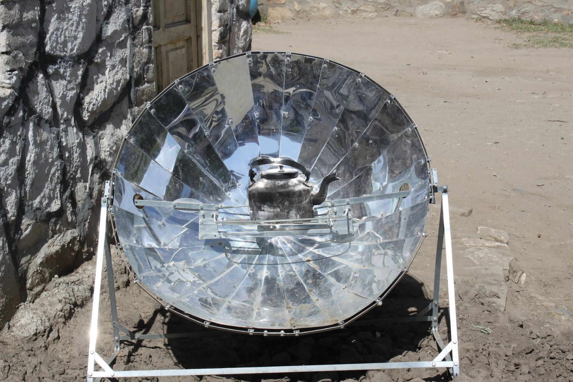 Zdjęcie przedstawia wielkie połyskujące zwierciadło kuliste wklęsłe z umieszczonym w ognisku czajnikiem. Zwierciadło ma białe trójkątne nogi. W tle znajduje się kamienna ściana, podłoże to żwir i piasek.