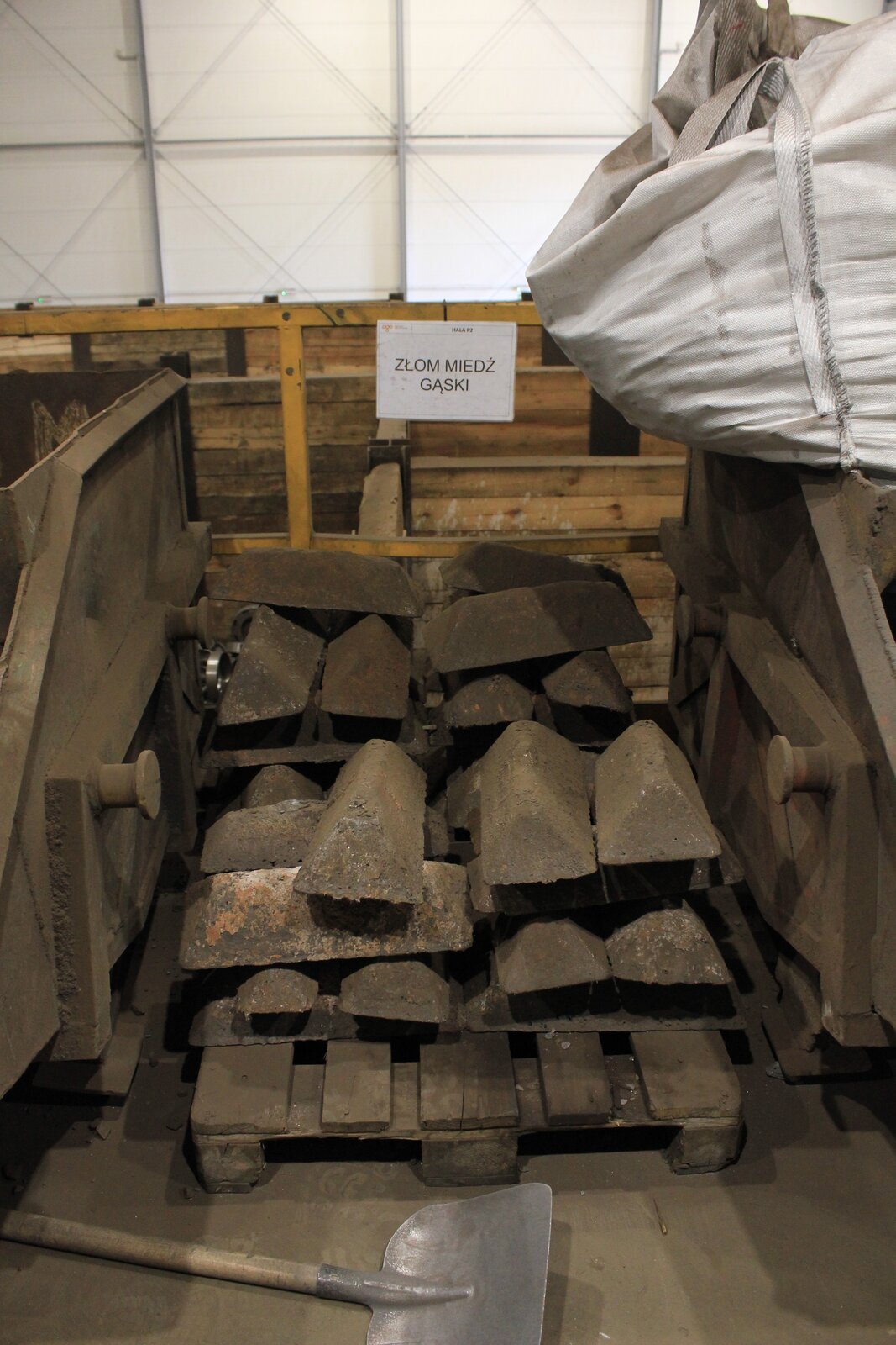 Na zdjęciu widoczne są gąski czystego metalu technicznego – miedź. Gąski przypominają budową sztabki. Leżą one na palecie. Po obu stronach są małe, otwarte kontenery na złom. Na kontenerze z prawej strony leży big bag. Przed paletą położona jest łopata. Za paletą zawieszona jest karteczka z napisem. ZŁOM MIEDŹ GĄSKI.