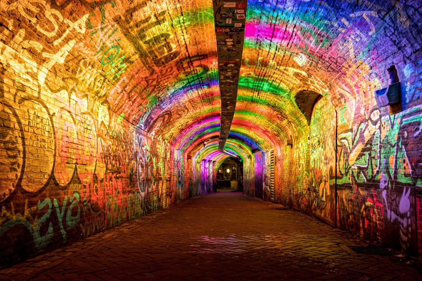 Zdjęcie przedstawia ściany tunelu/przejścia podziemnego, pokryte napisami i rysunkami. Napisy są różnych wielkości i mają różne kroje liter. Malowane są sprayami [czytaj: sprejami]. Mienią się kolorami od zimnych, chłodnych, poprzez gorące do ciepłych. Jak tapeta  pokrywają całą płaszczyznę ścian i sklepienia. 