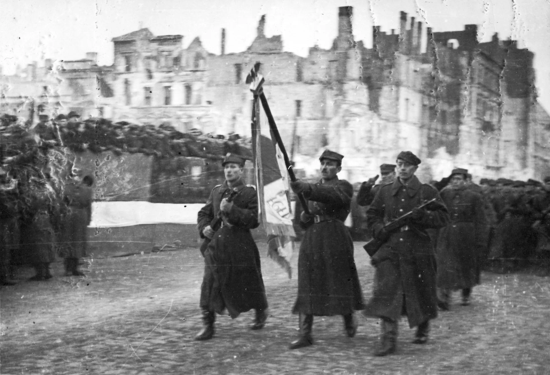 Kliknij, aby powiększyć Zdjęcie. Ulicą maszerują żołnierze. Ci, na przedzie niosą chorągiew i trzymają karabiny. Za nimi ciągnie się sznur idących żołnierzy. W tle, przy ulicy stoją ludzie. Kamienice za nimi są zrujnowane. Wypalone okna, brak dachów.