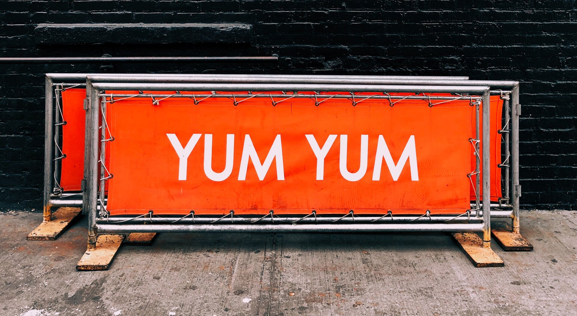 Zdjęcie przedstawia przykładowe banery drukowane. Widoczne są trzy prostokątne banery zamocowane na stalowych stelażach. W całości widoczny jest tylko pierwszy baner – na czerwonym tle widoczny jest duży napis z białych liter YUM YUM. Stelaże stoją na drewnianych podstawkach na betonowym podłożu. W tle czarna ściana.